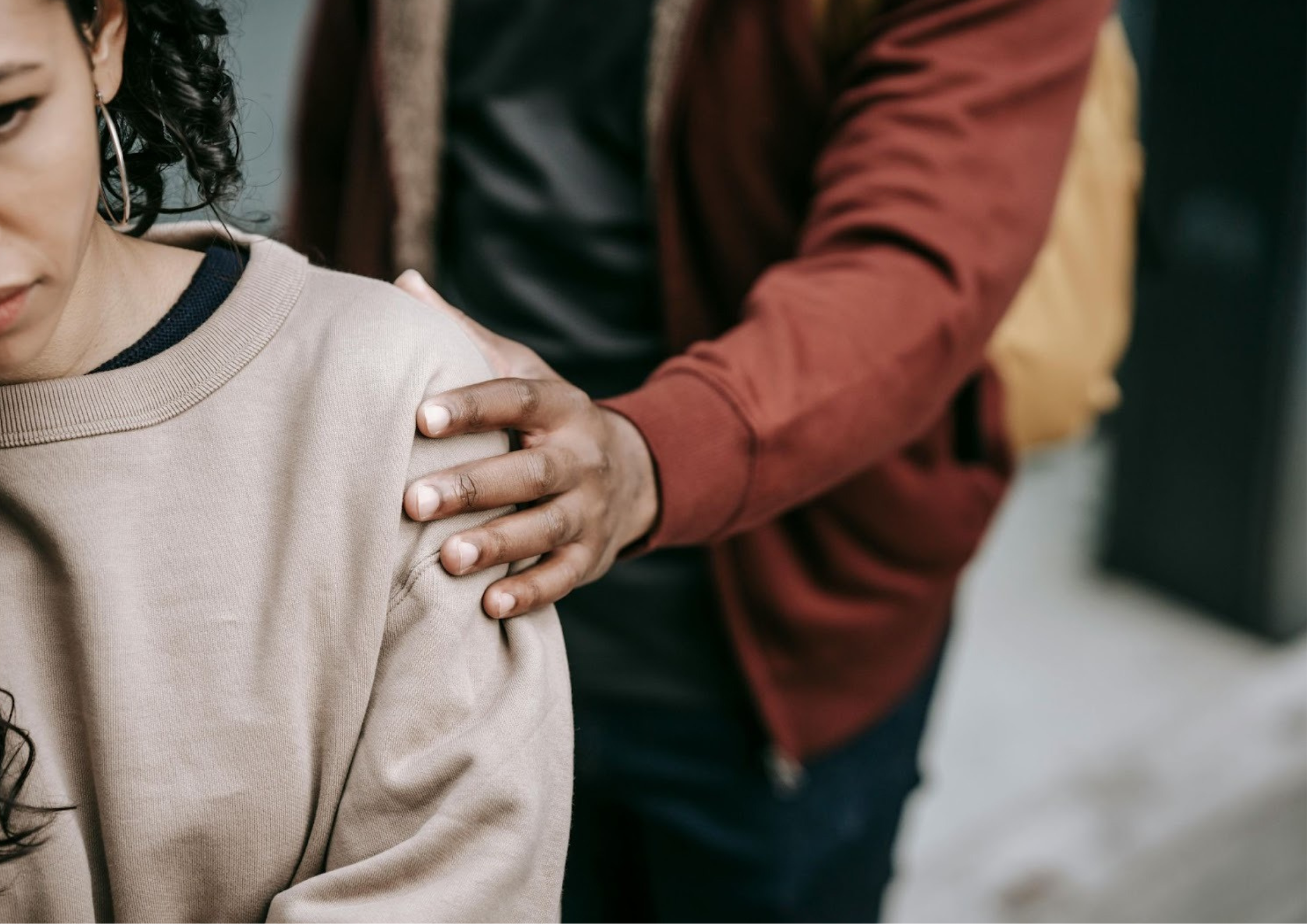 A close-up of a concerned woman looking down while another person gently places a supportive hand on her shoulder, suggesting emotional support during a difficult moment.