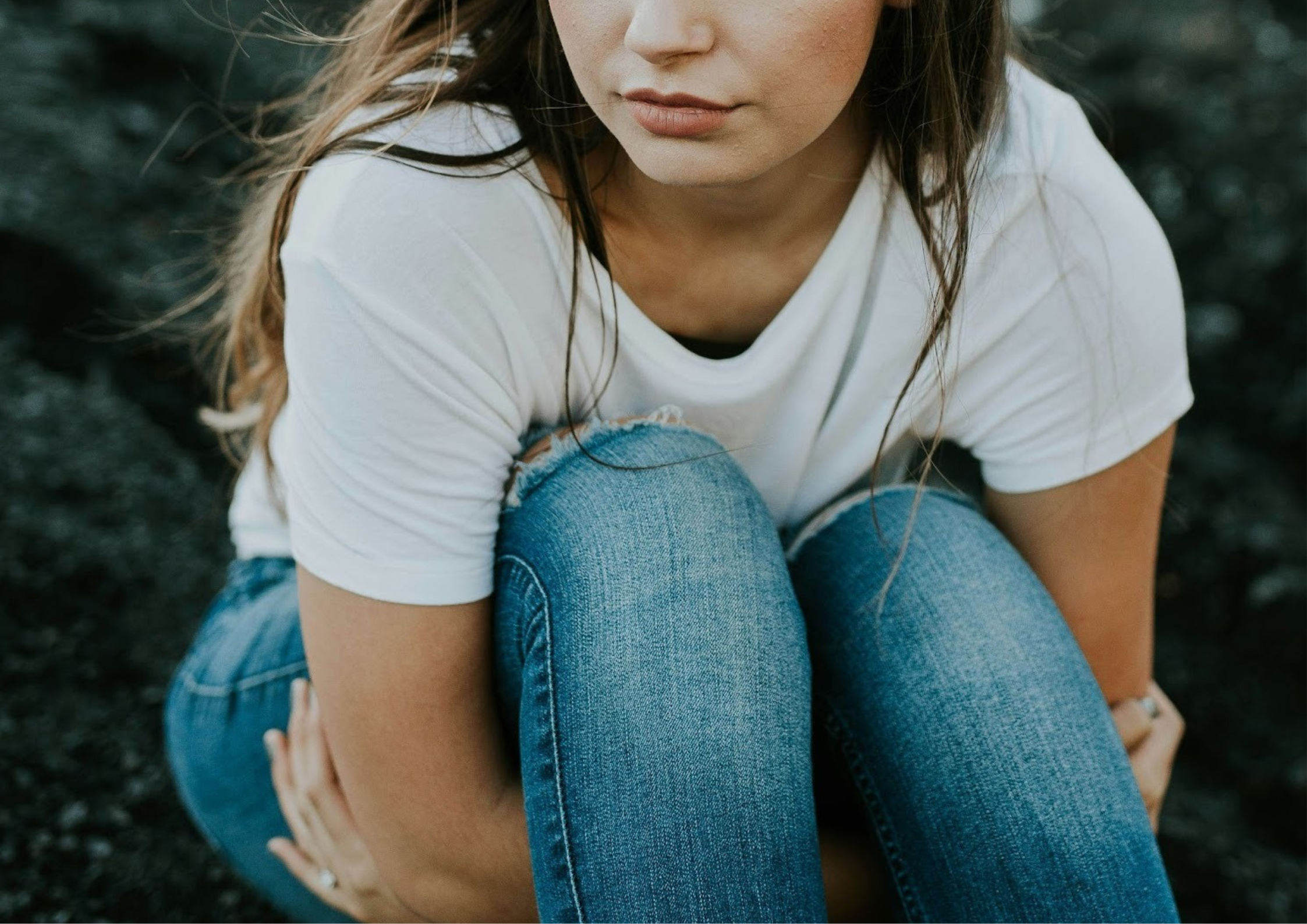 A close-up of a young woman sitting outdoors with her knees pulled to her chest, looking down with a pensive or anxious expression