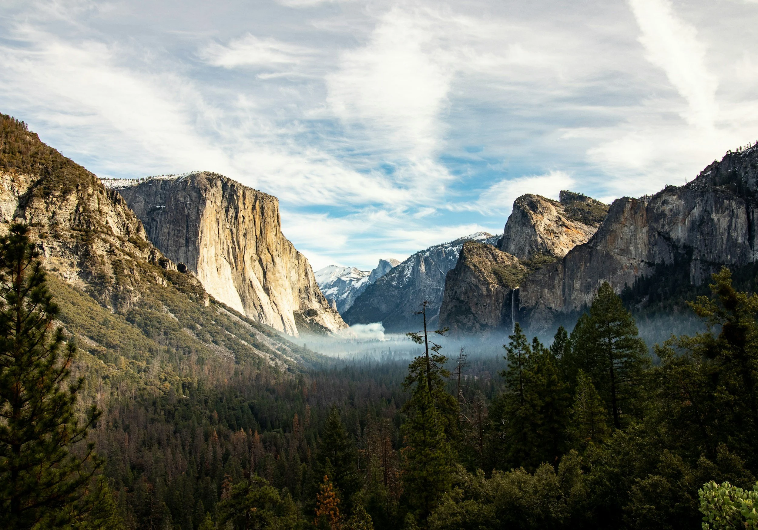 A scenic view of a valley with towering granite cliffs, lush green forest, and a partly cloudy sky.