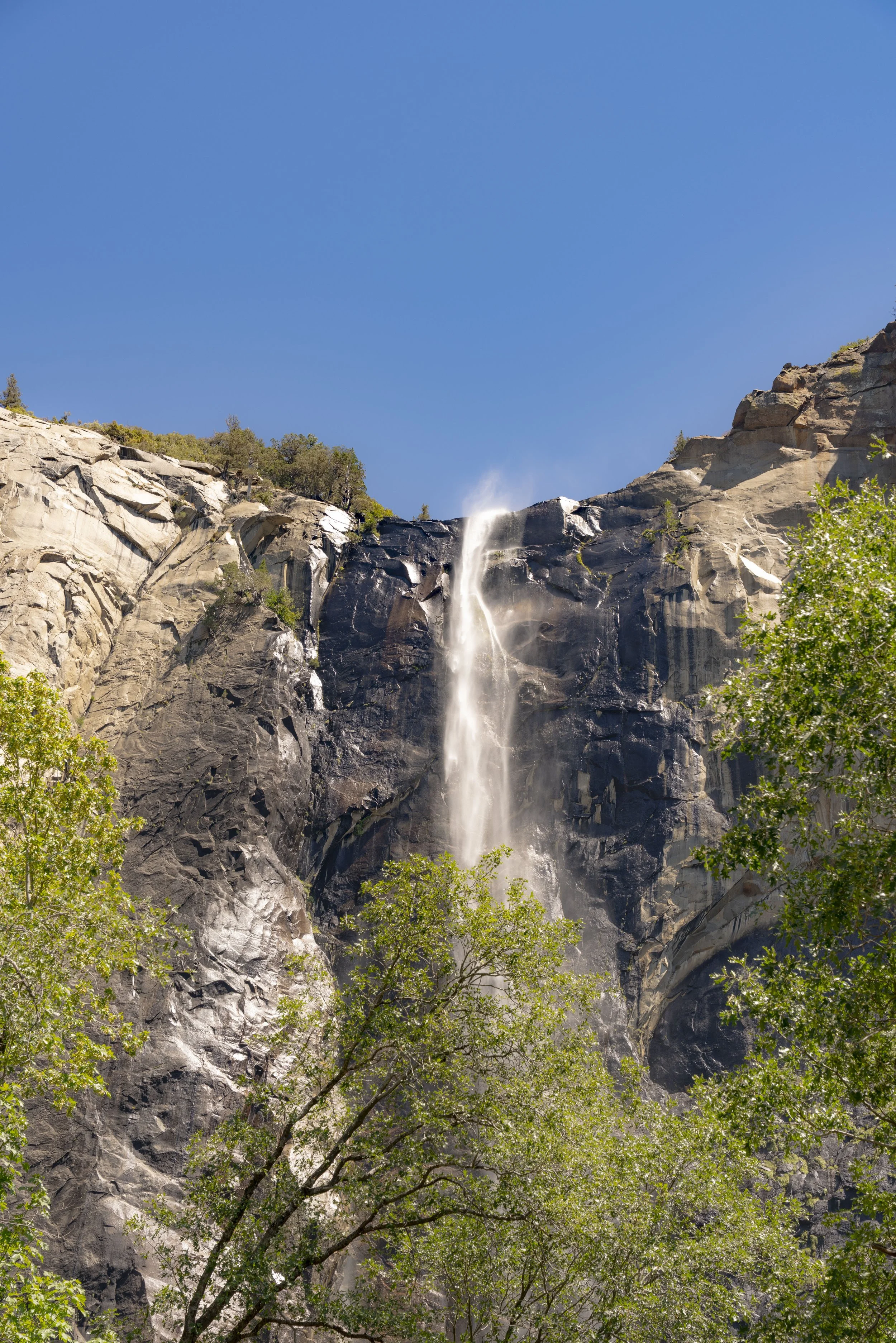 A tall waterfall flowing down the rugged face of a granite cliff surrounded by green trees under a clear blue sky.
