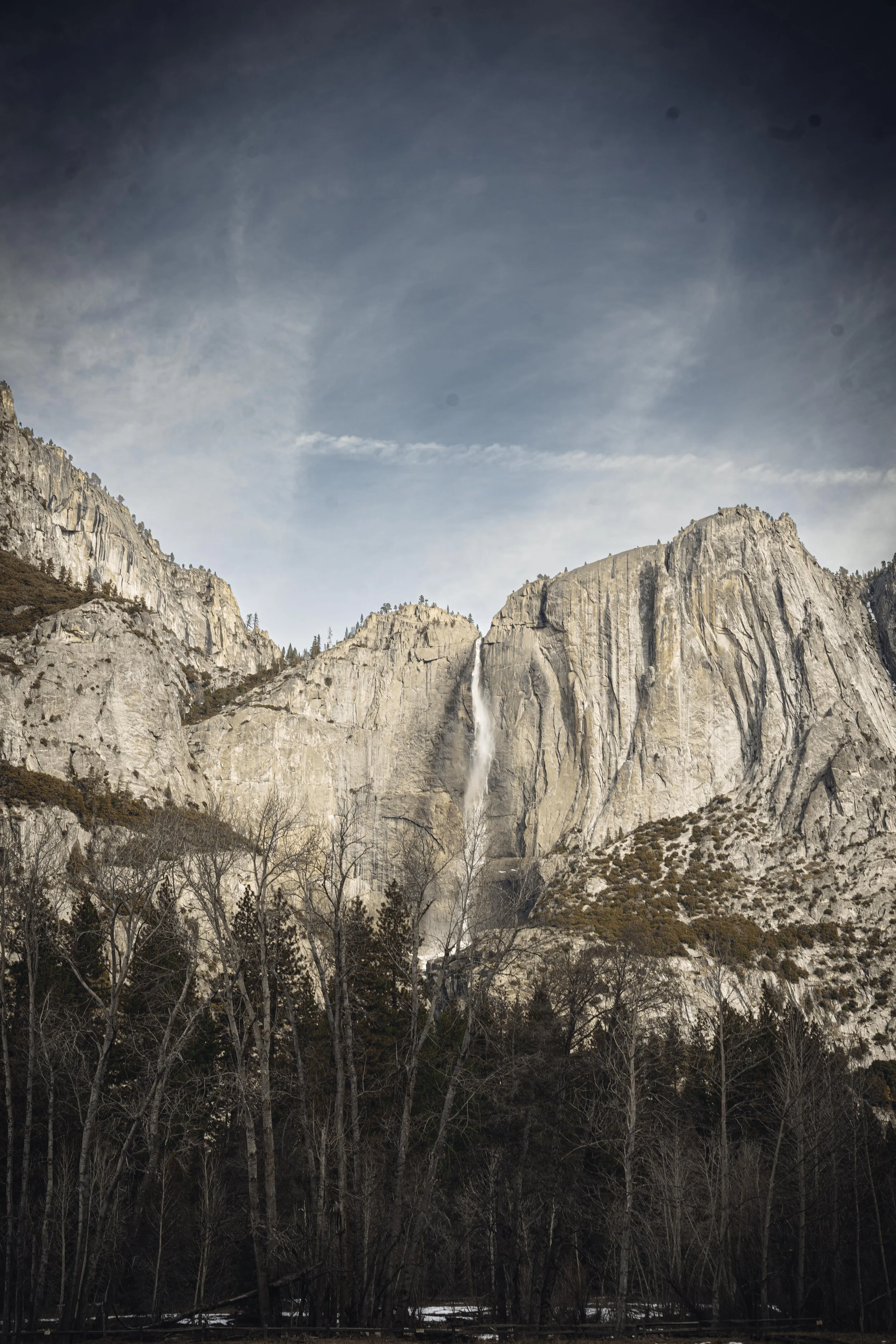 Scenic view of a tall waterfall cascading down granite cliffs, with leafless trees in the foreground and a cloudy sky overhead.