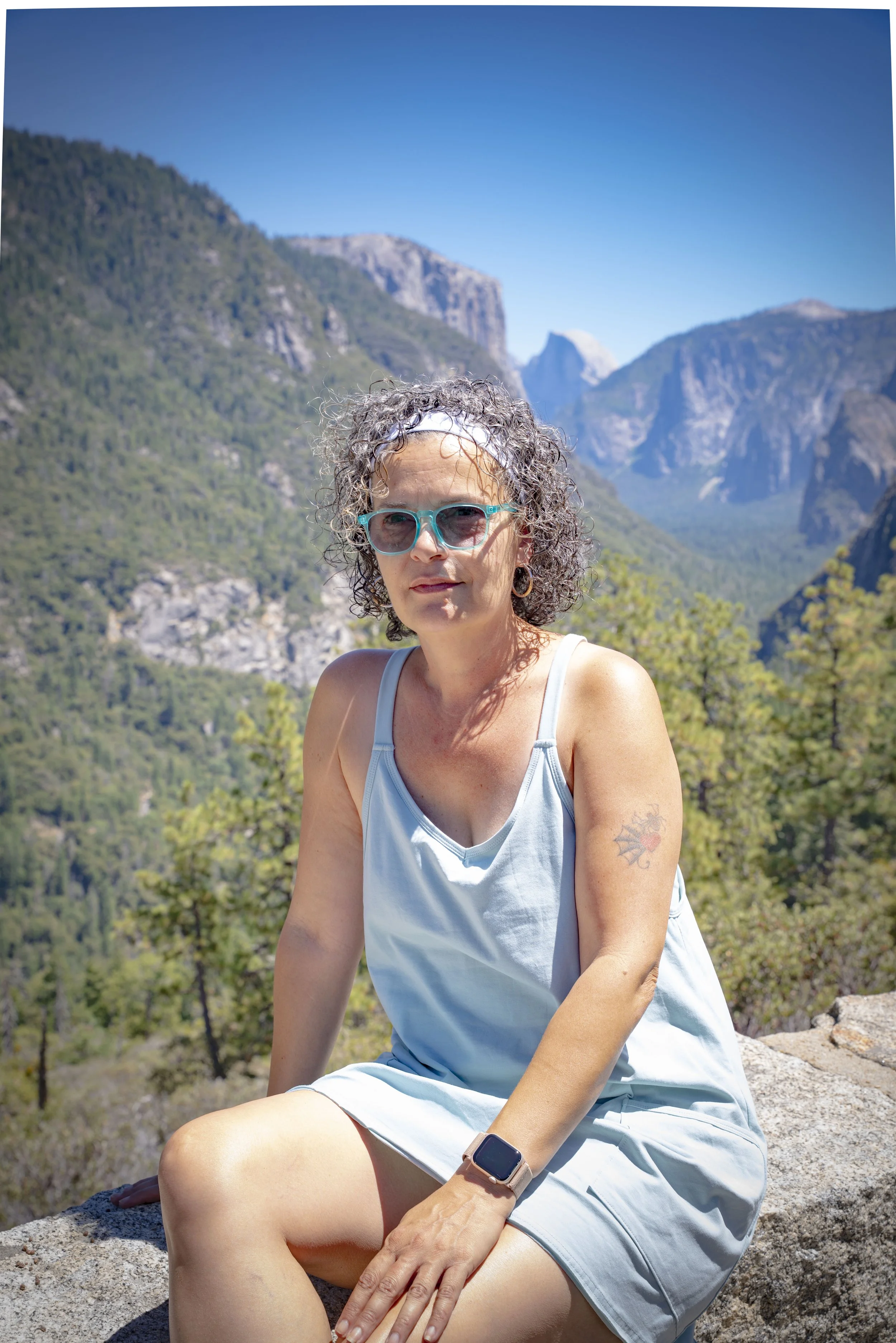 A woman with curly gray hair, sunglasses, and a bracelet sitting on a rock with a mountainous landscape in the background.
