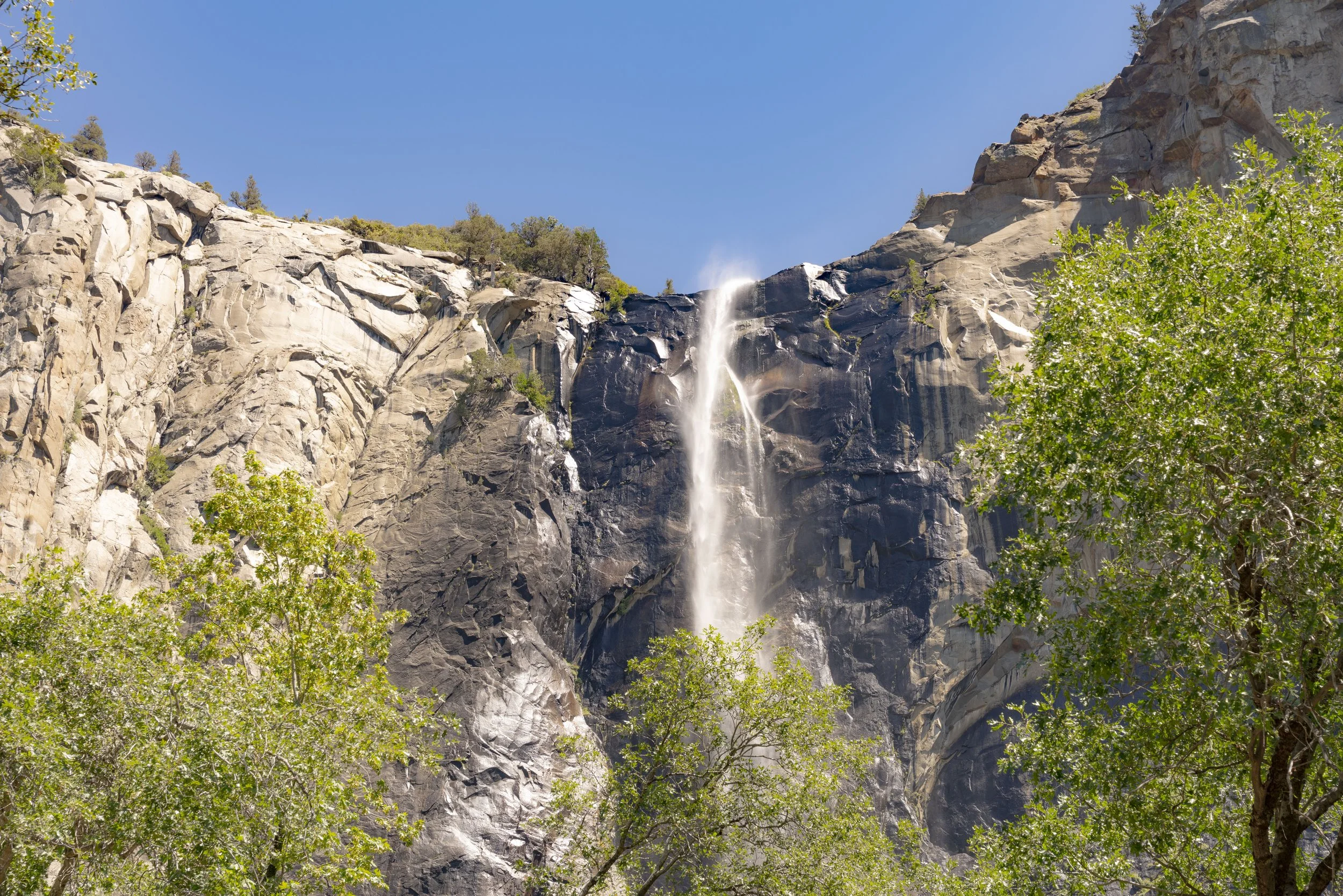 A tall waterfall cascading down a steep rocky cliff with some green trees in the foreground and a clear blue sky above.