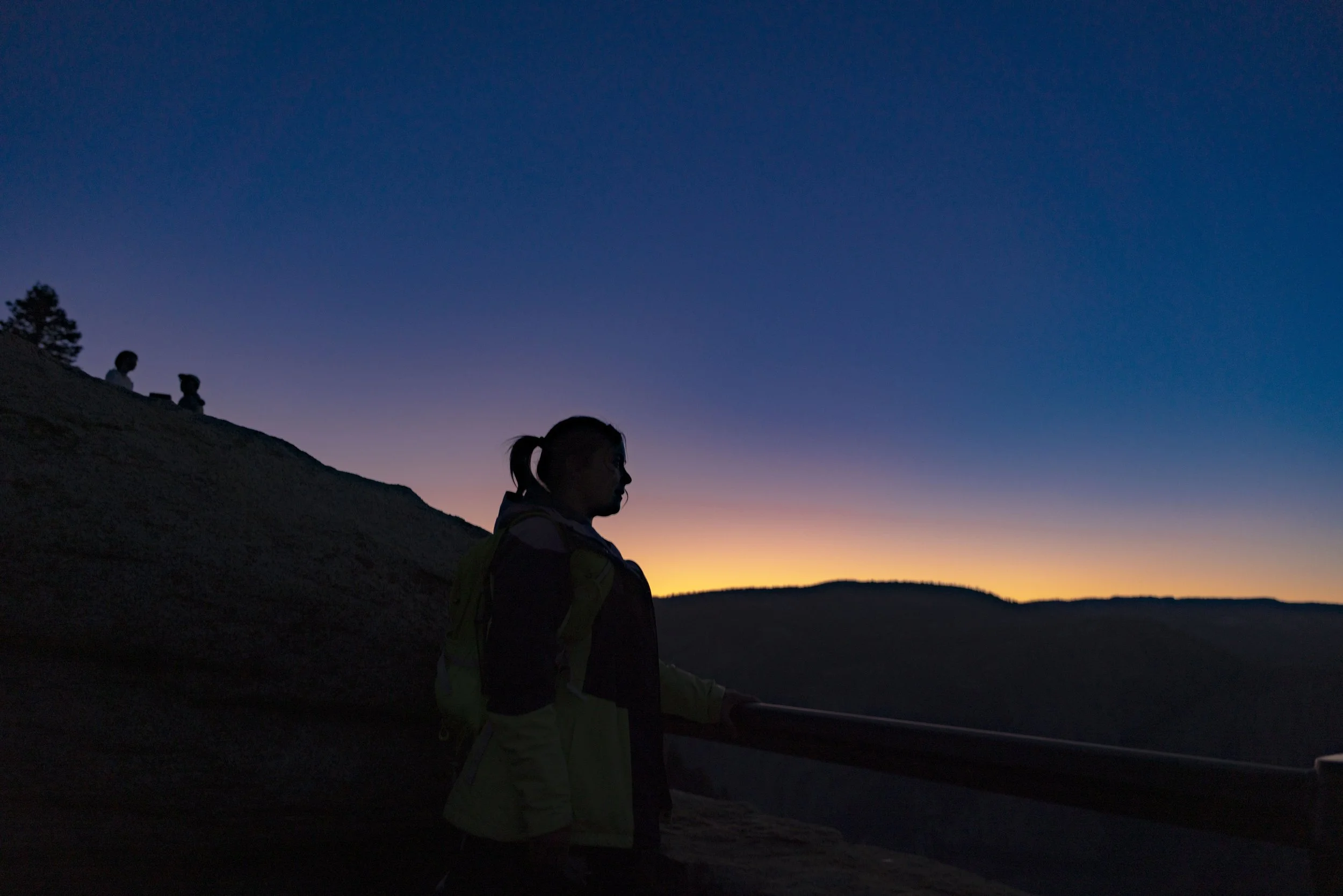 Silhouette of a person standing by a railing during dusk, with a dark mountain slope and a colorful sunset sky in the background.