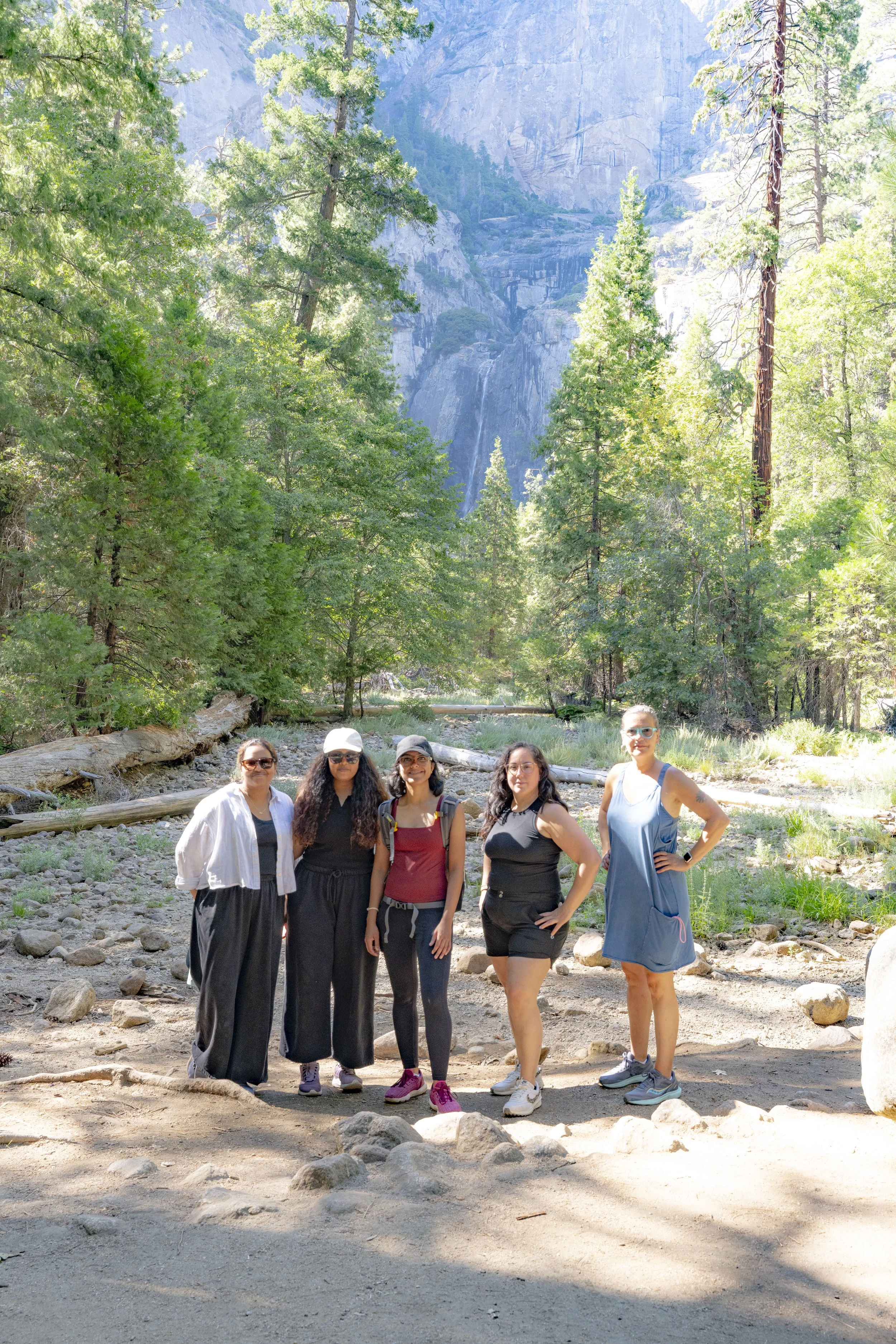 Five women standing together outdoors in a forested area with tall trees and a mountain in the background, dressed in casual hiking attire.