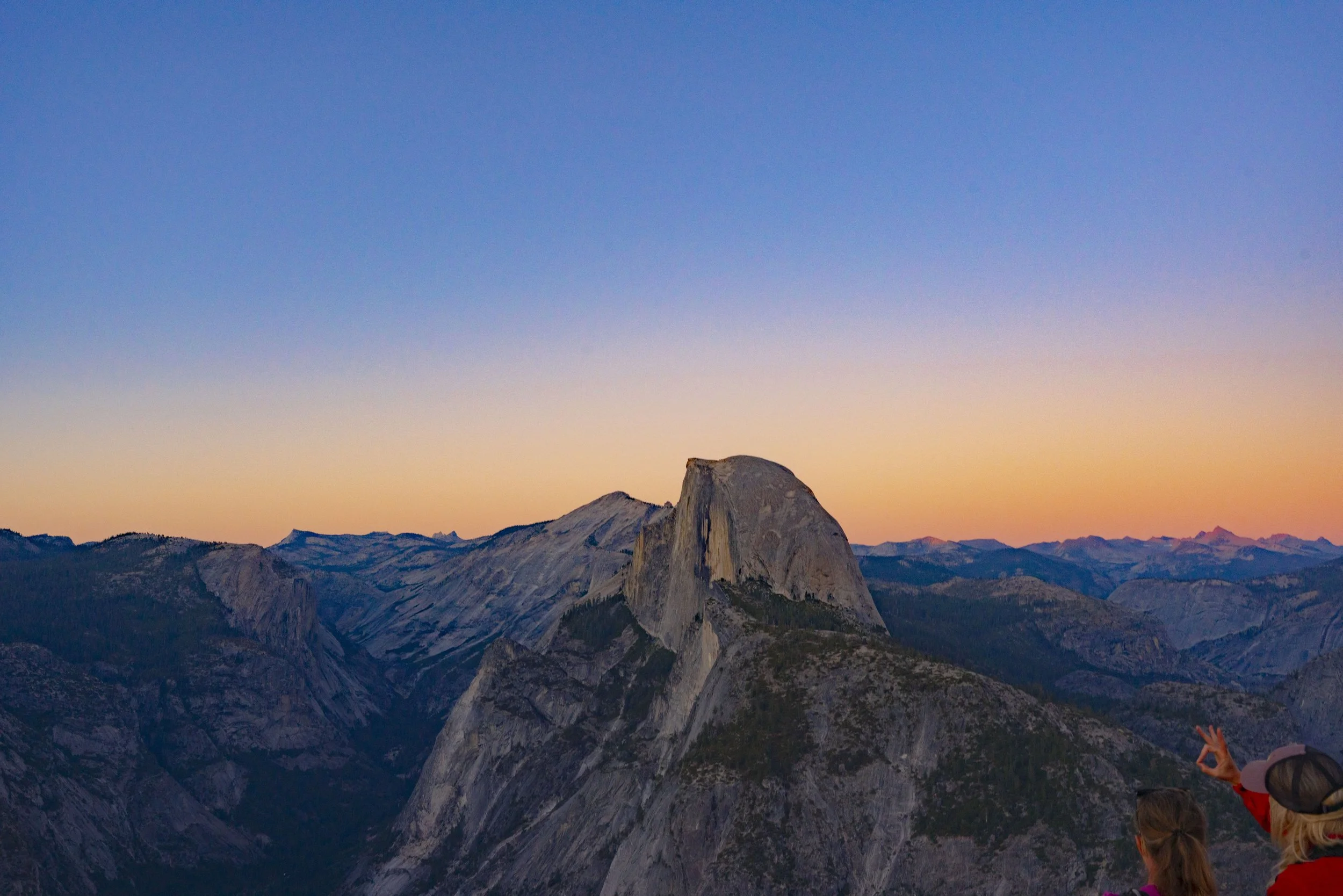 View of Yosemite National Park with Half Dome at sunset, with two people in the foreground, one making an OK gesture.