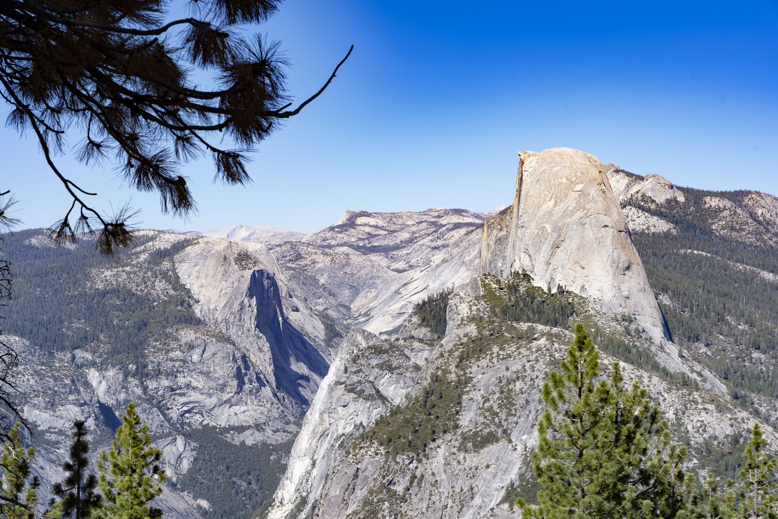 A scenic view of a mountain range with notable granite formations, pine trees, and a clear blue sky.