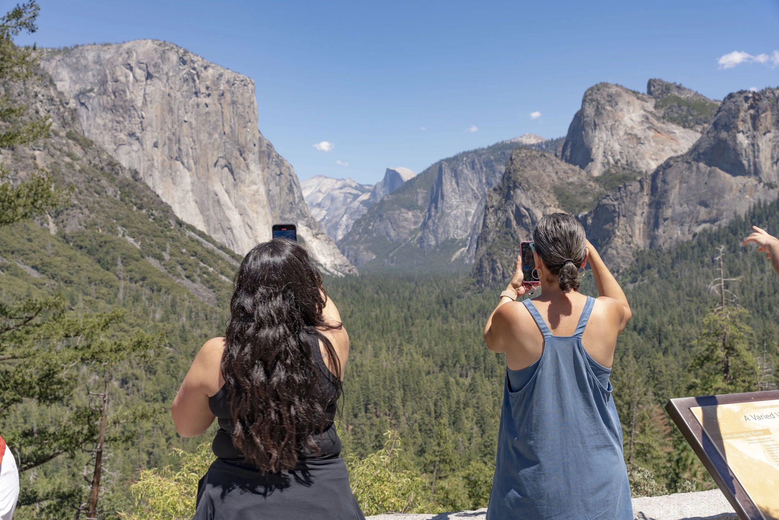Two women with long dark hair, one in a black top and the other in a blue dress, taking photos of a mountain landscape with their smartphones at a scenic lookout in a national park.