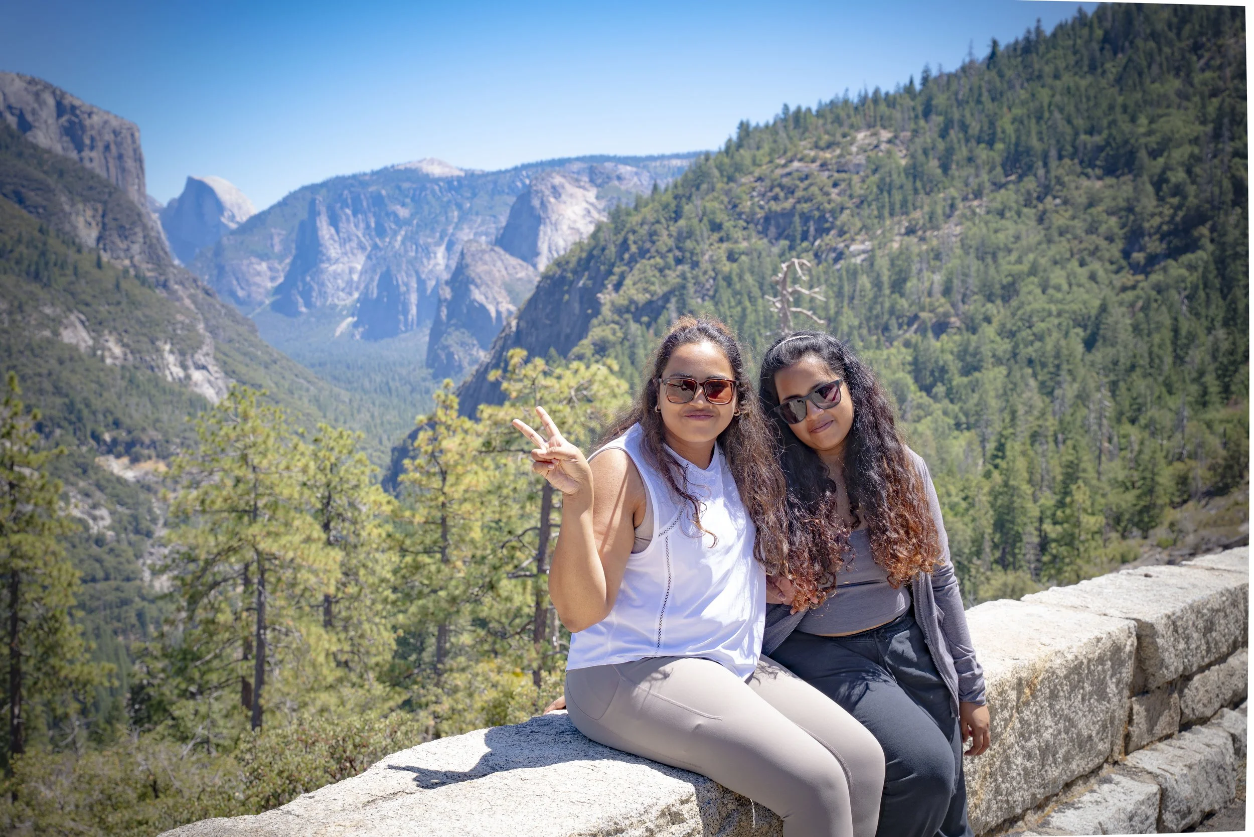 Two women sitting on a stone wall amidst a mountainous forest landscape, with trees, mountains, and blue sky in the background, one making a peace sign.