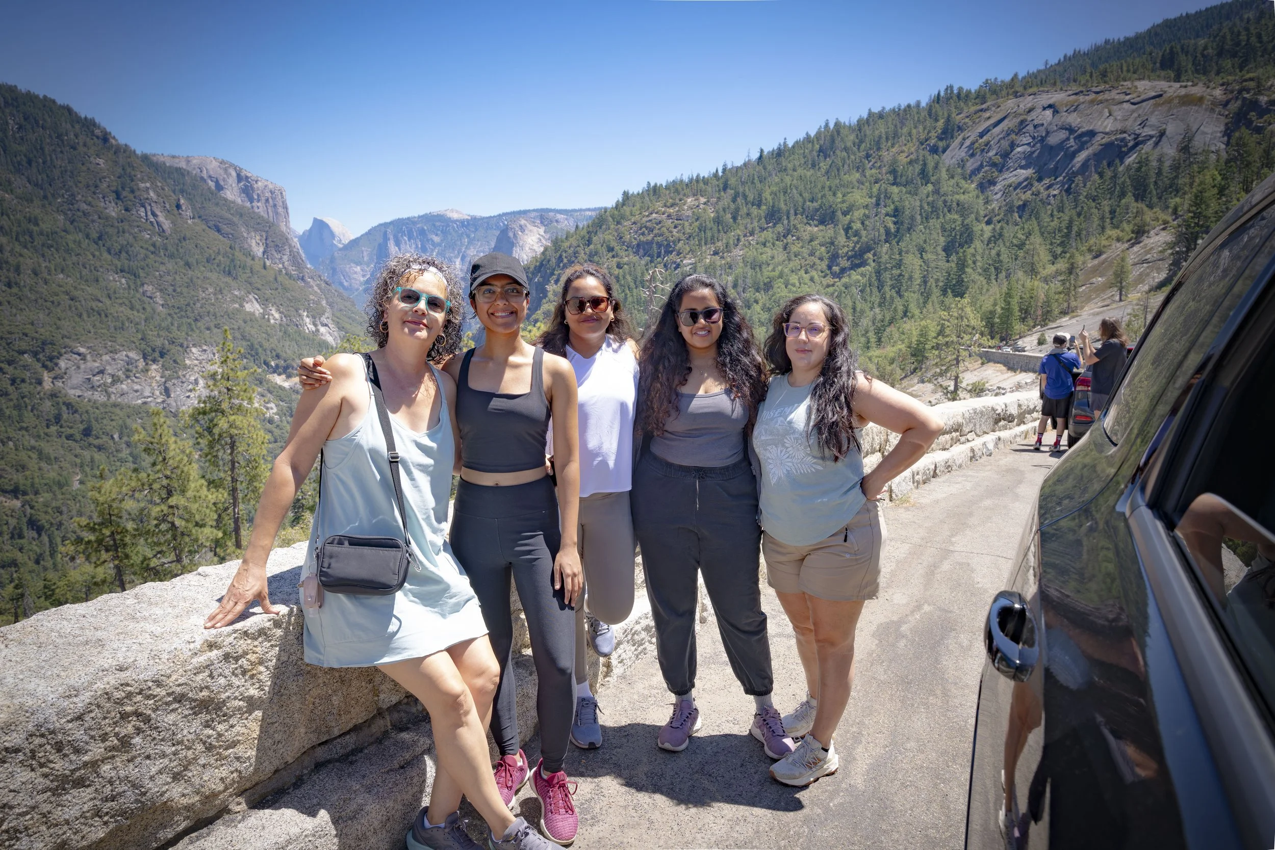 Five women posing together outdoors in a mountainous area on a sunny day, with lush green forests and cliffs in the background.