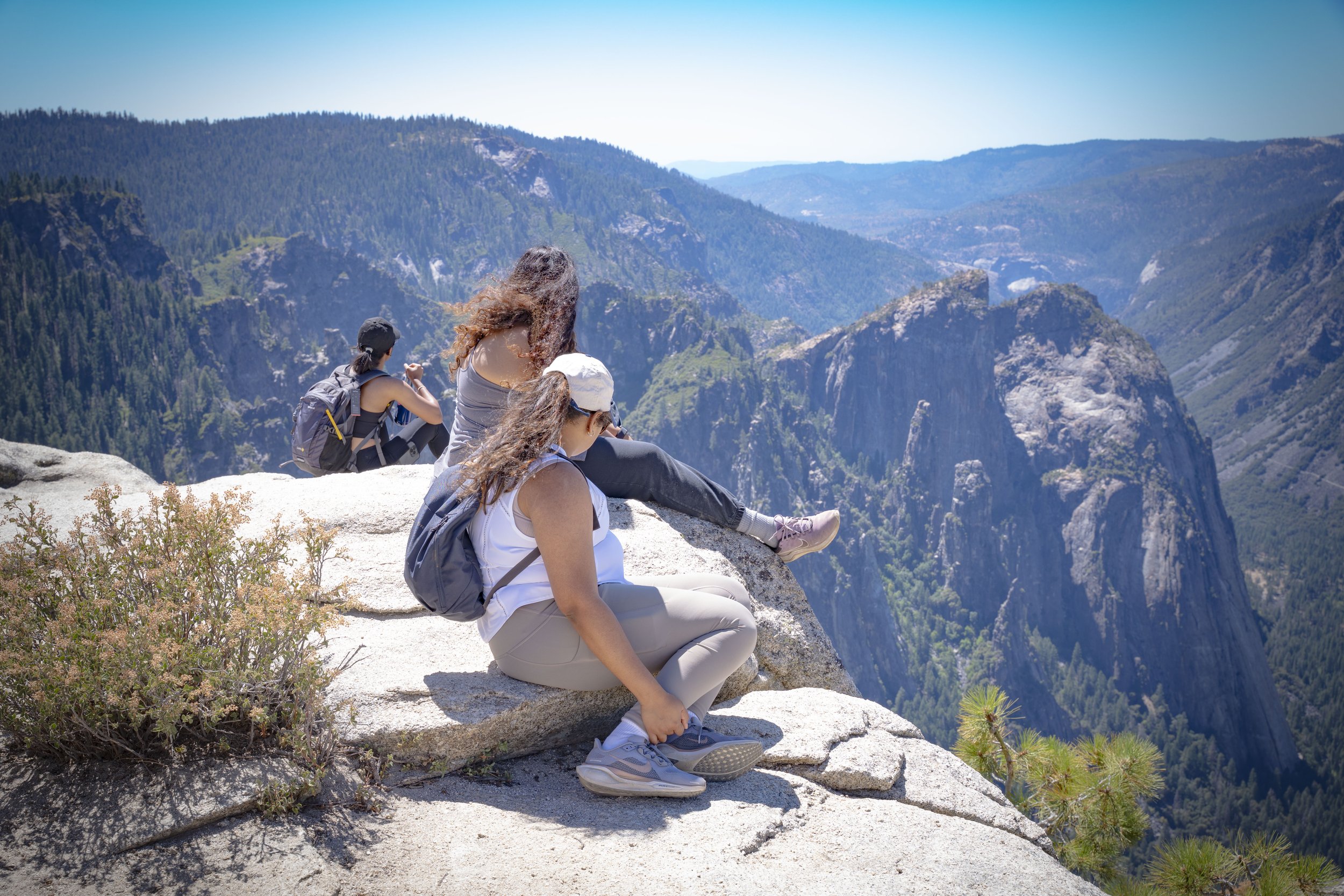 Three women sitting on a rocky ledge overlooking a mountainous valley, with forested cliffs and peaks in the background on a sunny day.
