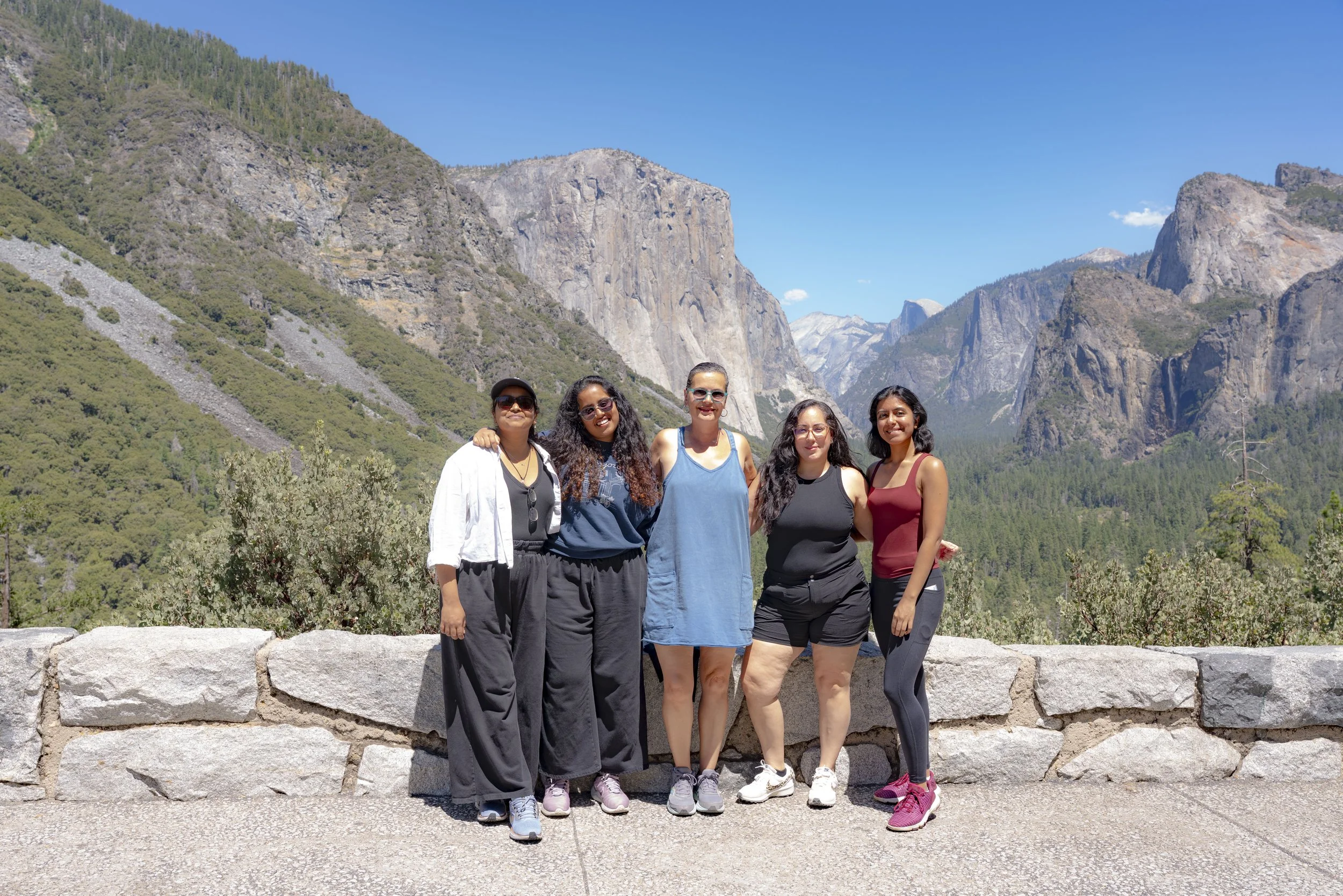 Five women standing together in front of a mountain landscape at Yosemite National Park, with large rock formations and lush green trees surrounding them.