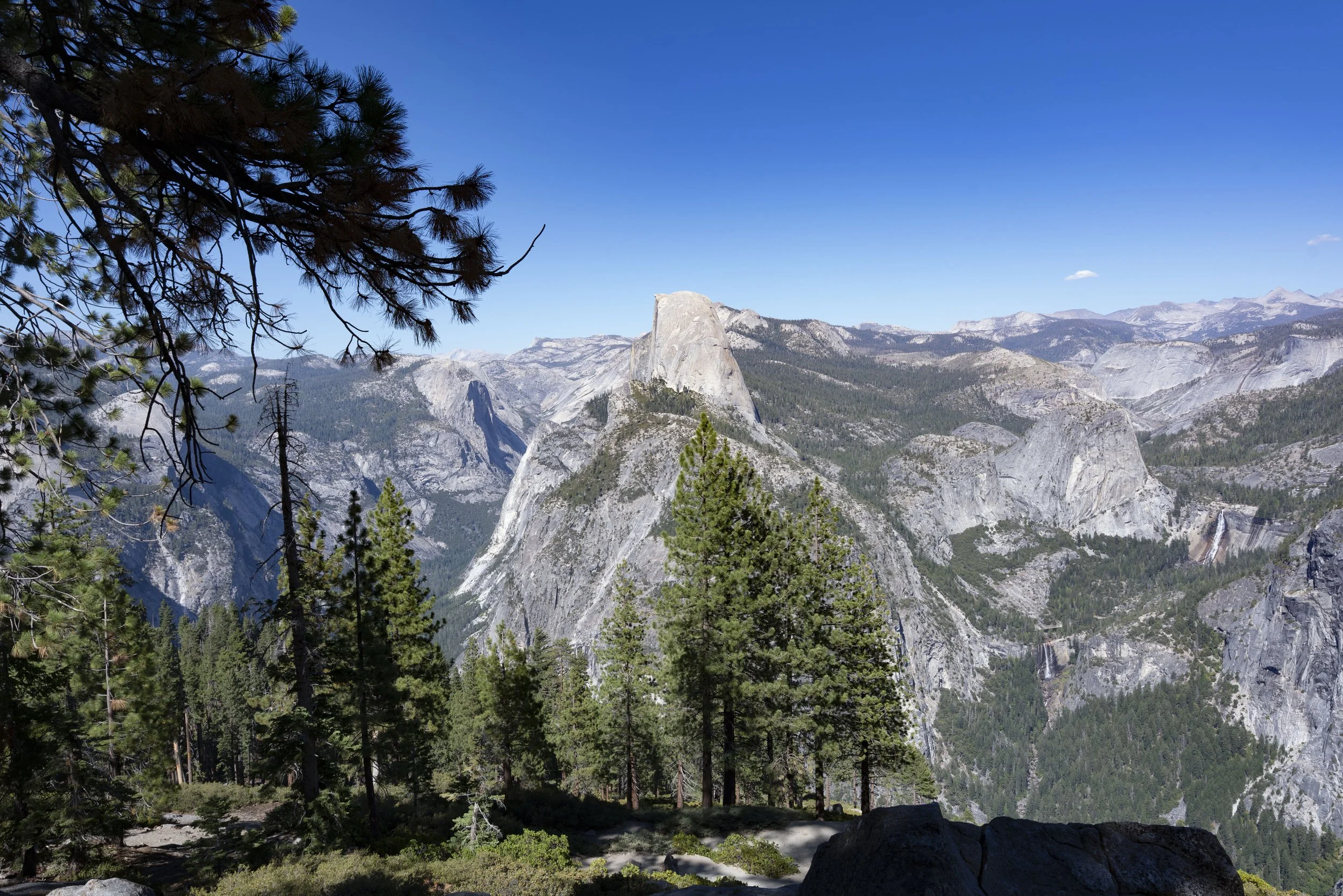 Scenic view of a mountain range with a prominent granite formation, surrounded by pine trees and a clear blue sky.