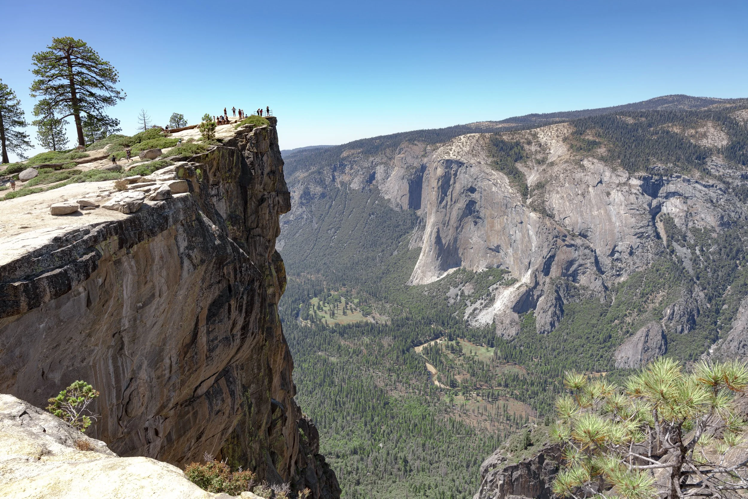 A panoramic view of a deep mountain valley with steep cliffs and rocky formations, and a group of tourists standing on a flat rocky ledge at the top of the cliff, surrounded by pine trees and green forested mountains in the distance under a clear blu
