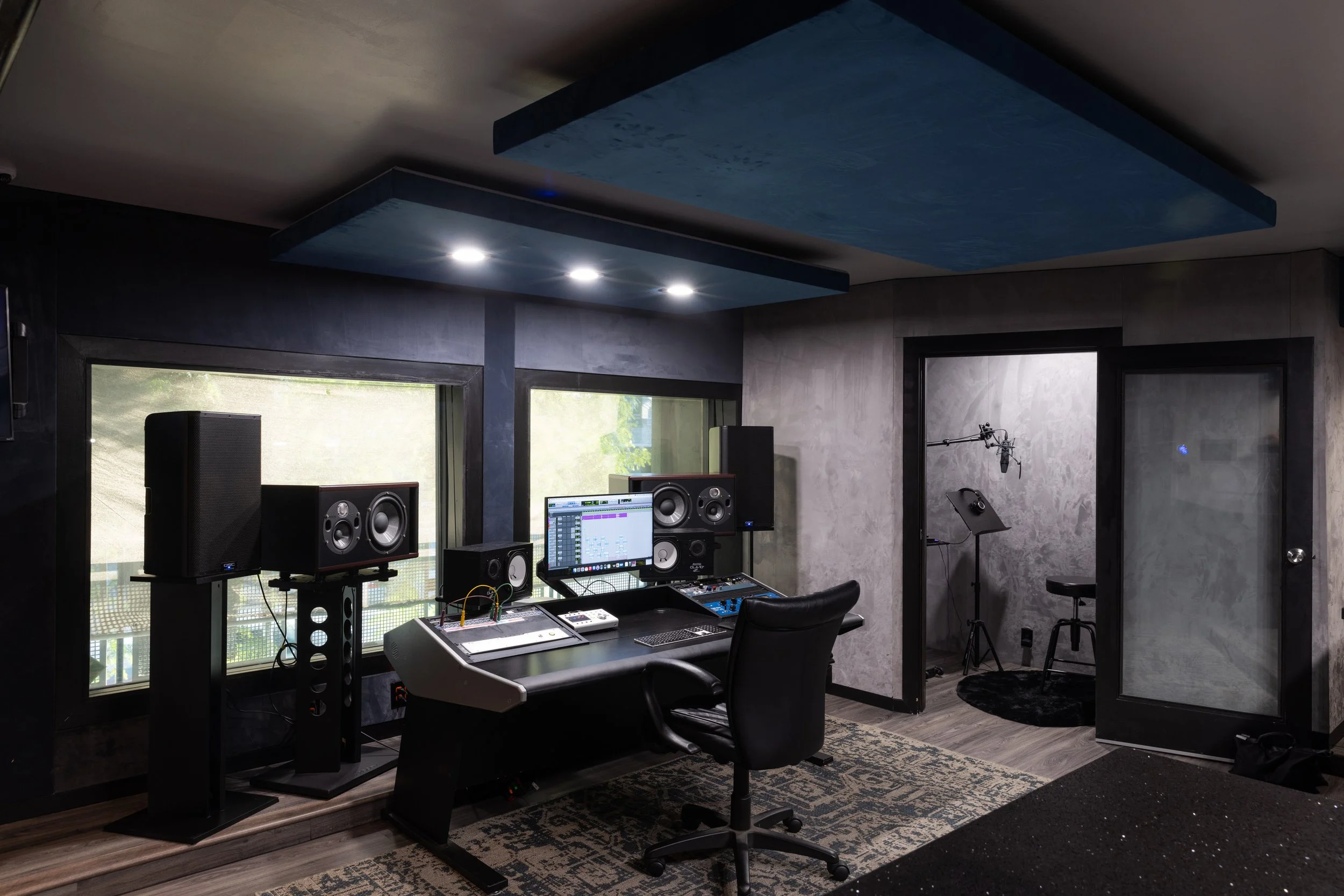 Modern music recording studio with mixing desk, large monitors, computer screen, and vocal booth with microphone setup.
