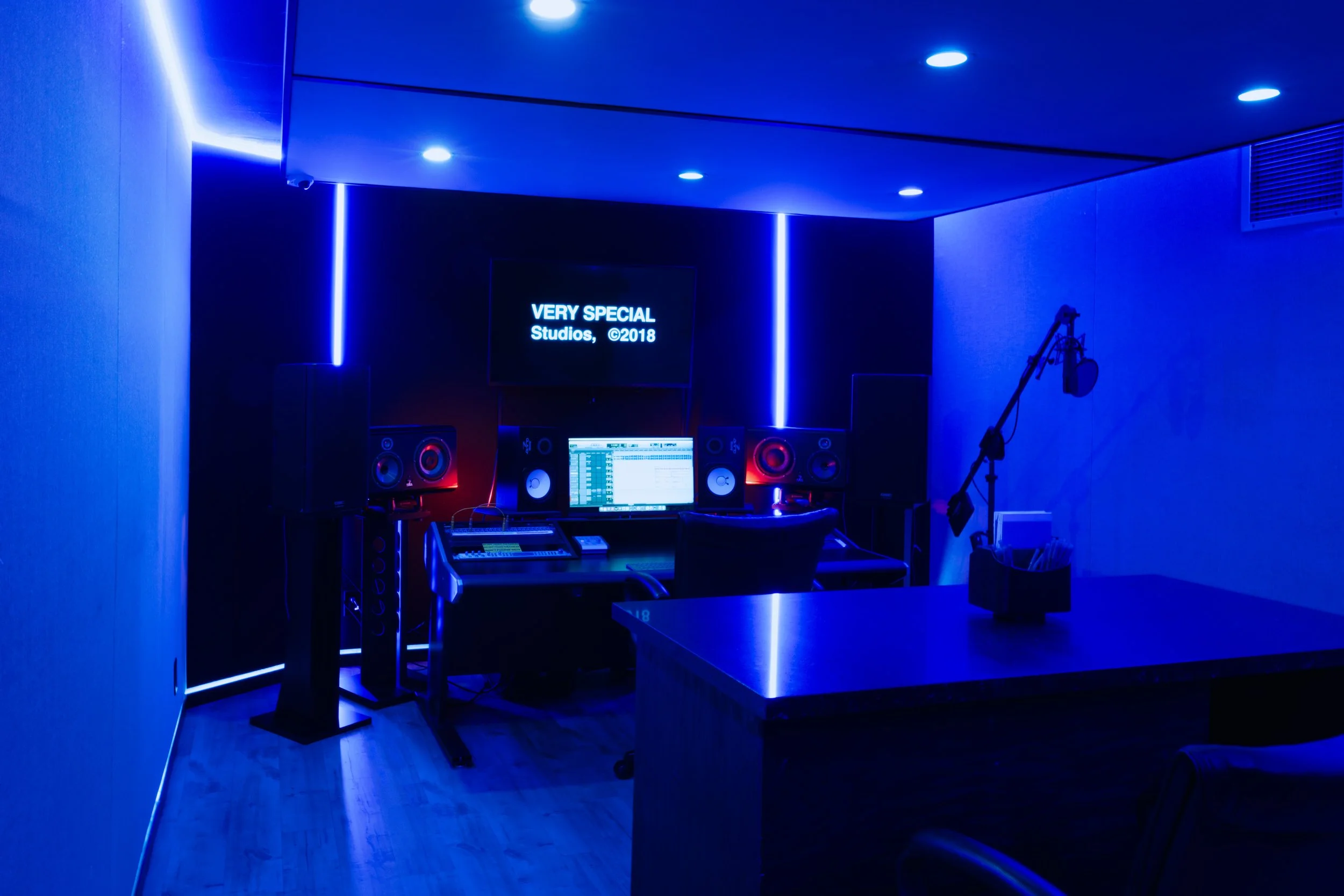 Modern recording studio with blue LED lighting, desk with computer monitor, speakers, microphone, and acoustic panels.