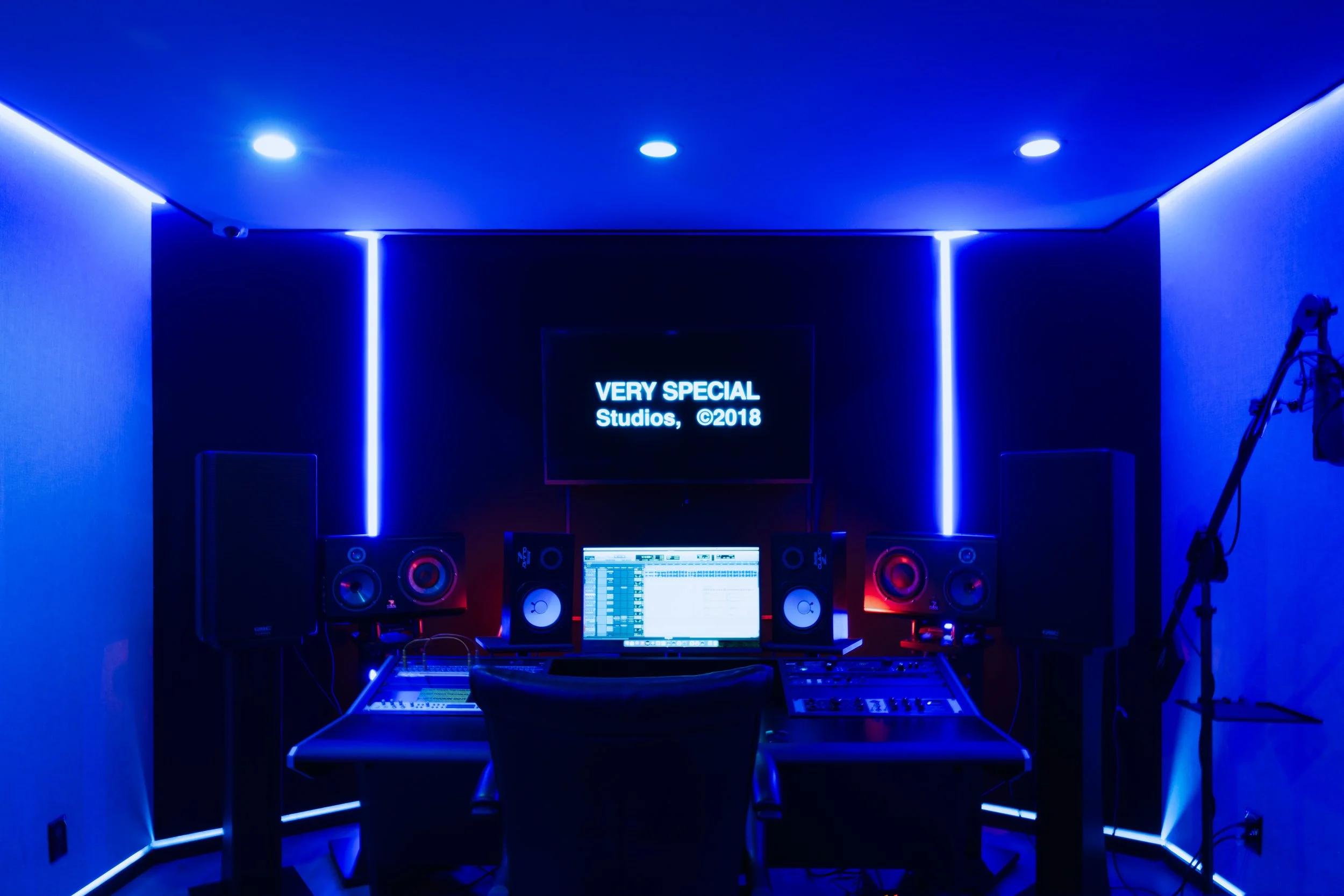 Interior of a music studio with blue ambient lighting, featuring two large speakers, a mixing console, a computer screen with audio software, and a microphone. A screen displays text: 'VERY SPECIAL Studios, ©2018.'