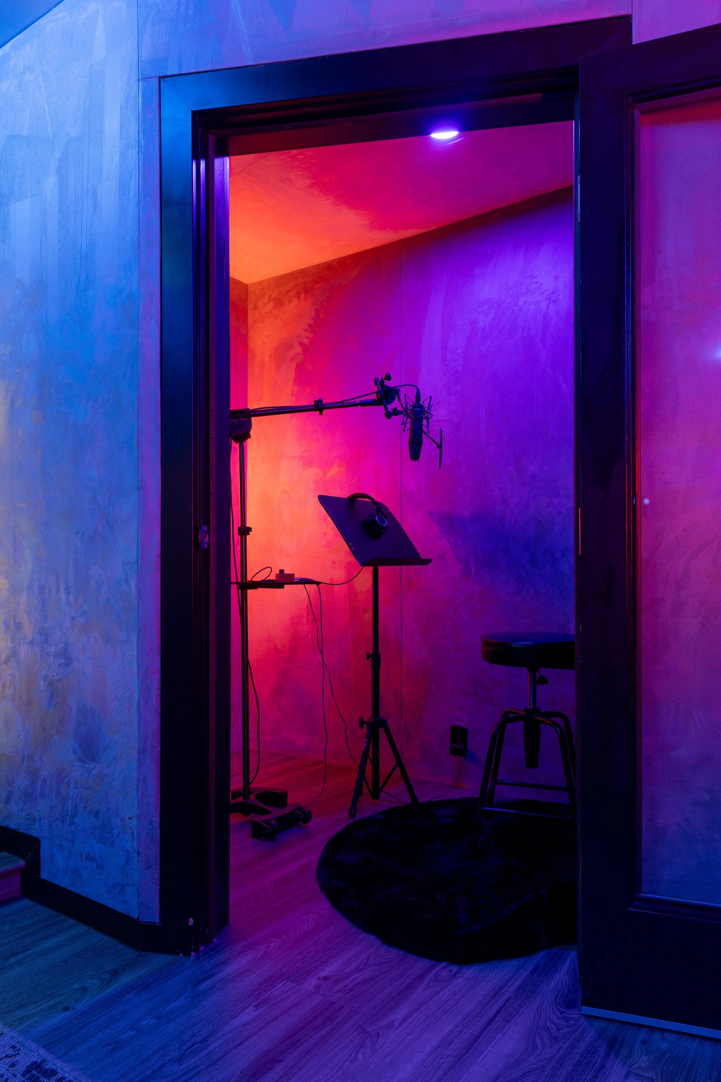 Recording booth with microphone, music stand, and stool under purple and orange lighting.