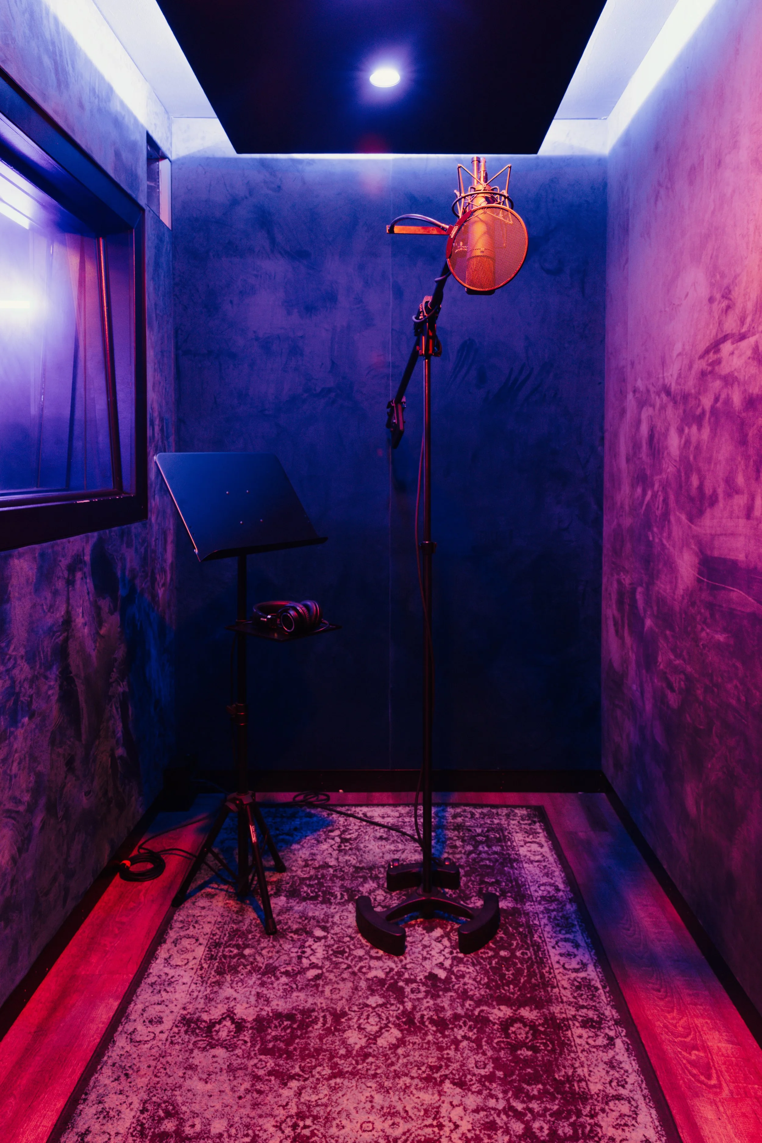 Empty recording studio booth with microphone, stand, headphones, music stand, and illuminated wall.