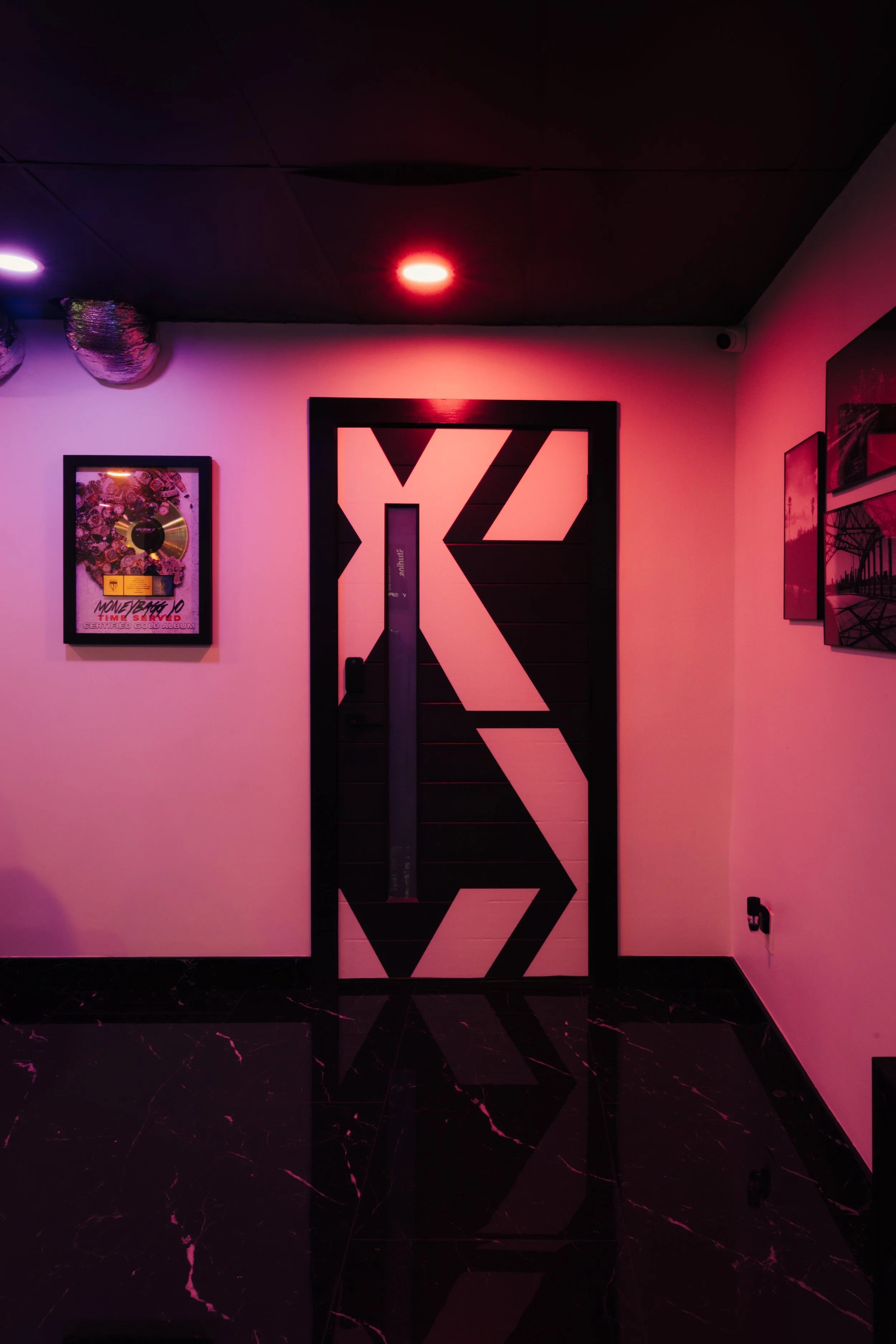 Interior with pink lighting, geometric patterned door, wall art including a framed record, and polished black floor.