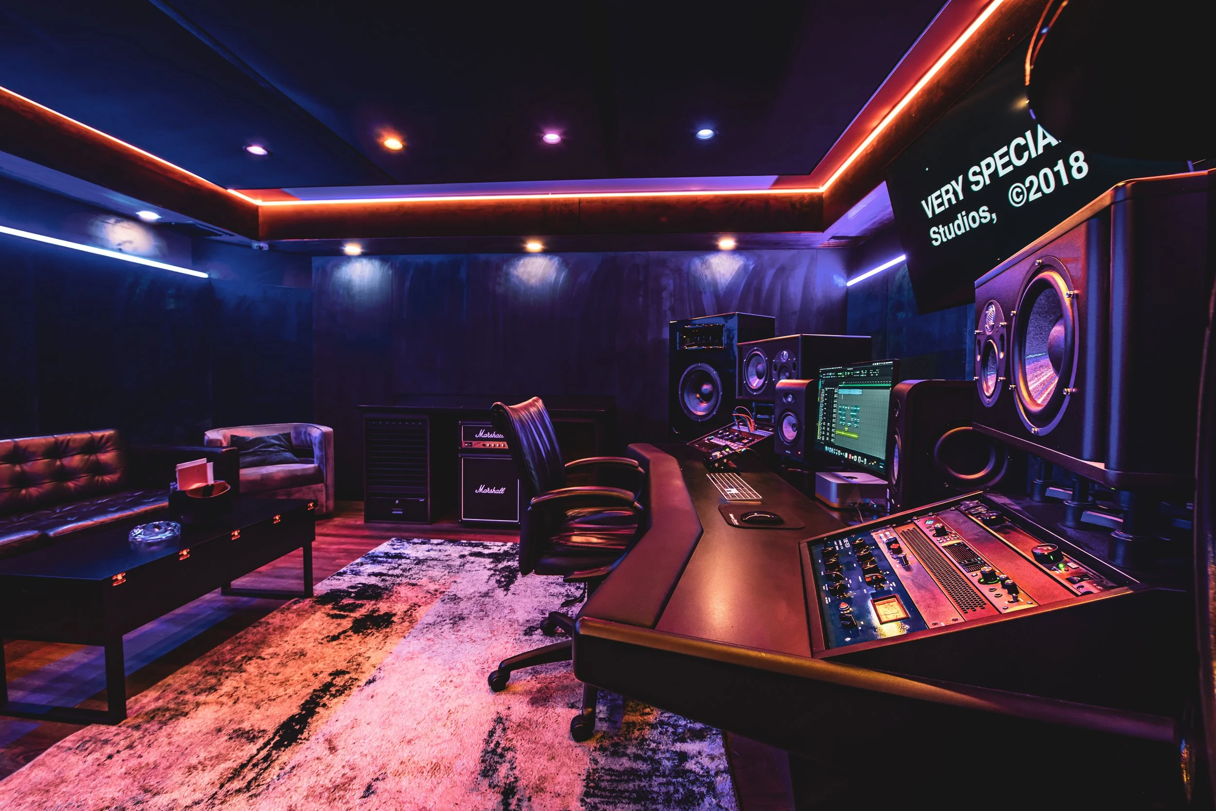 Recording studio room with audio equipment, monitors, speakers, mixing console, leather chairs, sofa, and dim lighting.