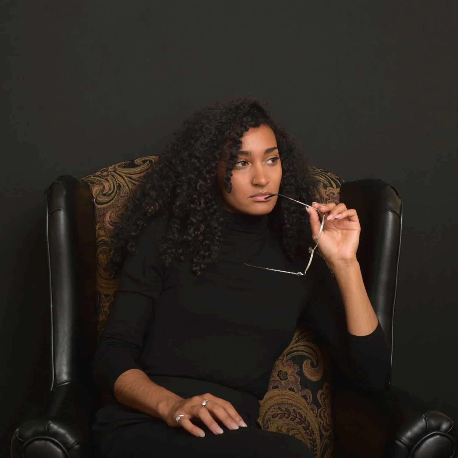 A portrait of MacKenzie (@zenkenzie) sitting in a black leather armchair against a black background, holding glasses near her lips, wearing a black turtleneck and silver jewelry.