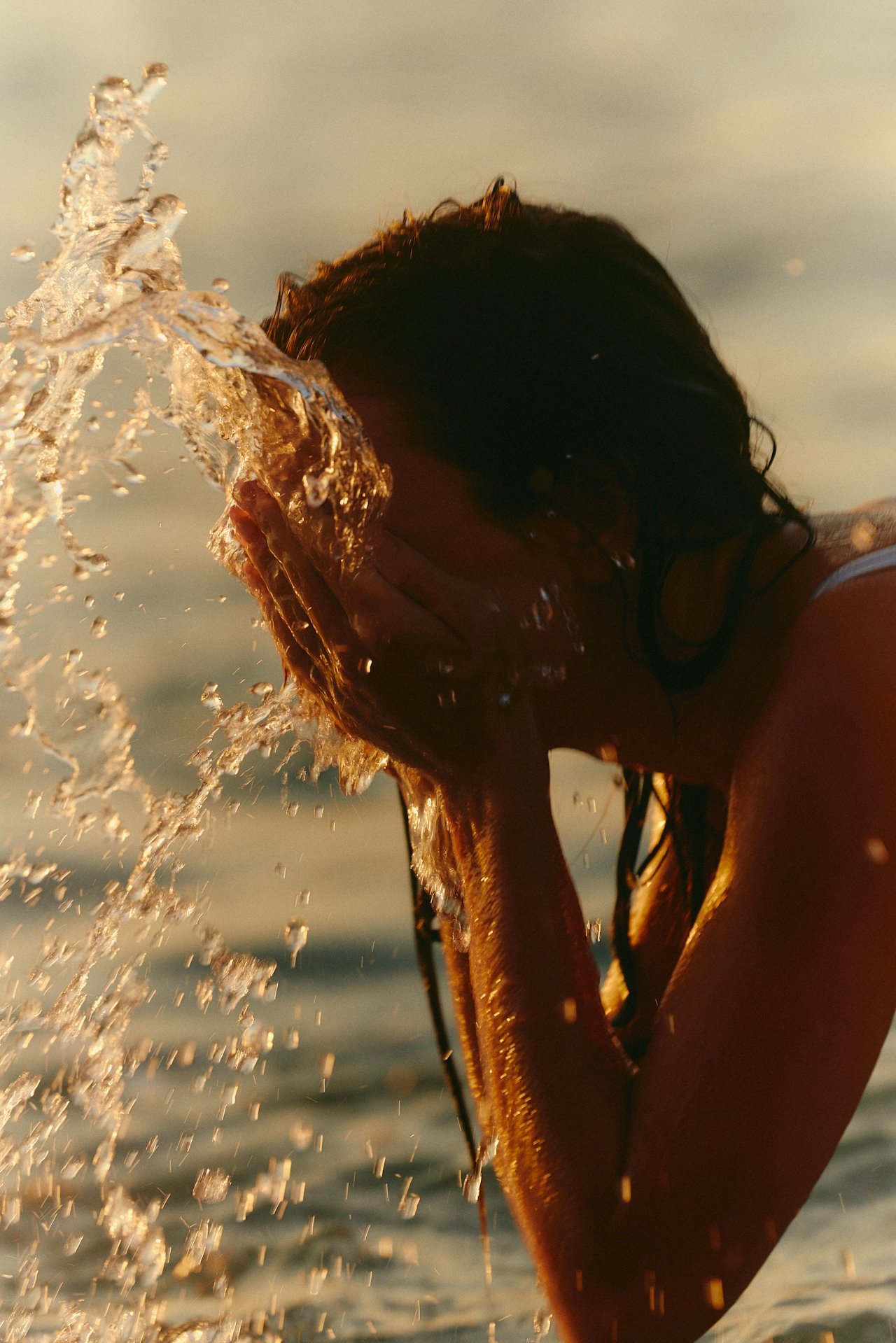 Person with wet hair splashing water at the beach during sunset.