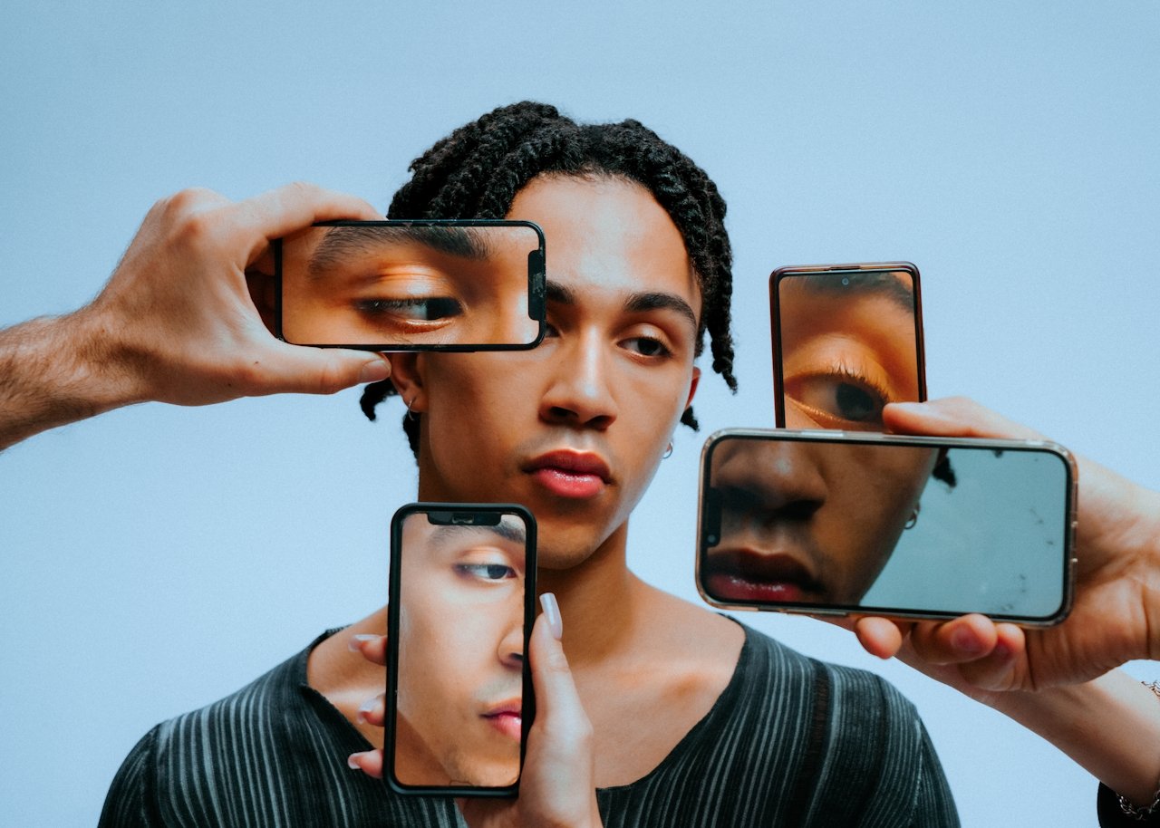 A person with short curly hair looks into multiple handheld mirrors and phone screens reflecting close-up views of their eye and face against a plain light blue background.