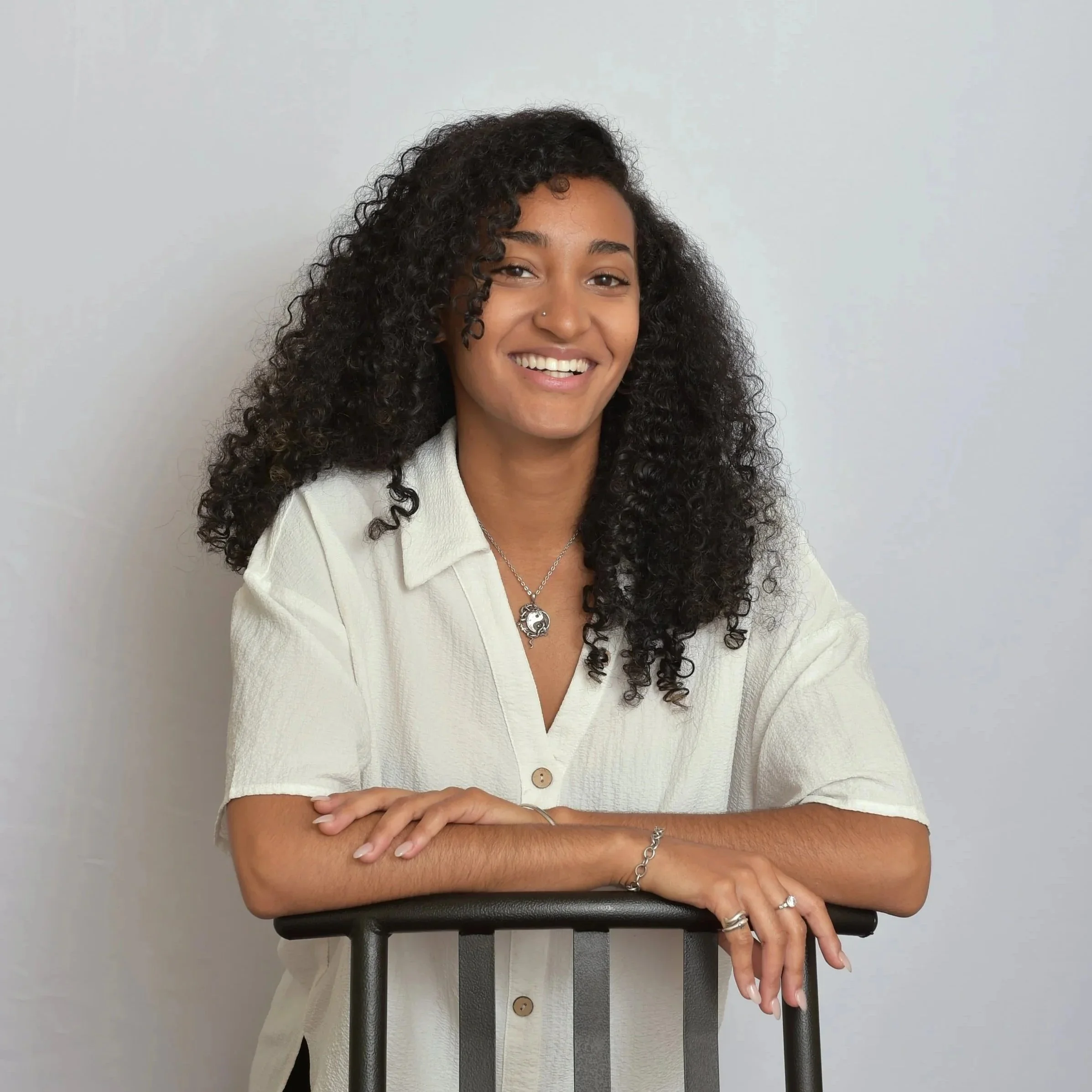 Portrait of a MacKenzie (@zenkenzie) wearing a white shirt and yin yang necklace, leaning on a chair against a plain white backdrop.