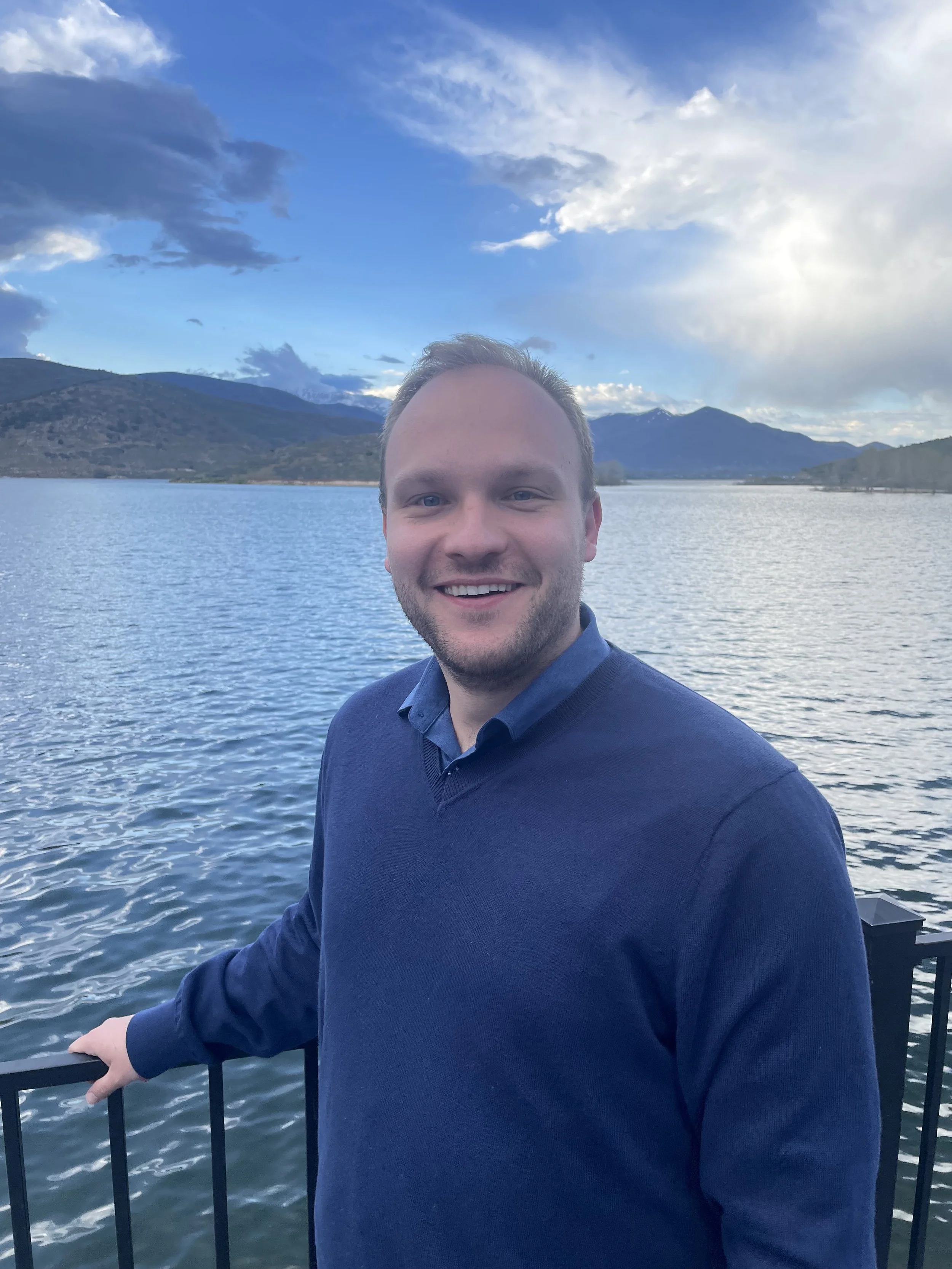 A smiling man in a navy blue sweater standing by a railing with a lake and mountain range in the background under a partly cloudy sky.