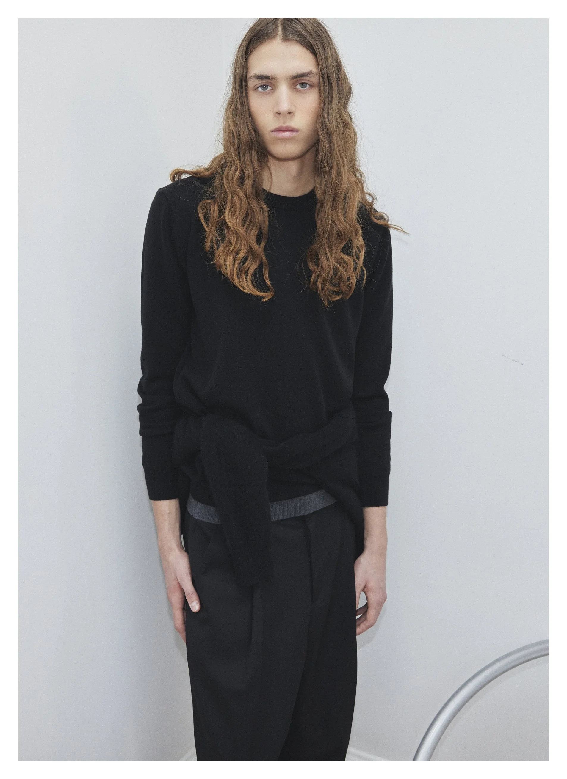 Jeune personne aux cheveux longs et ondulés, portant un pull noir et un pantalon noir, debout contre un mur blanc.