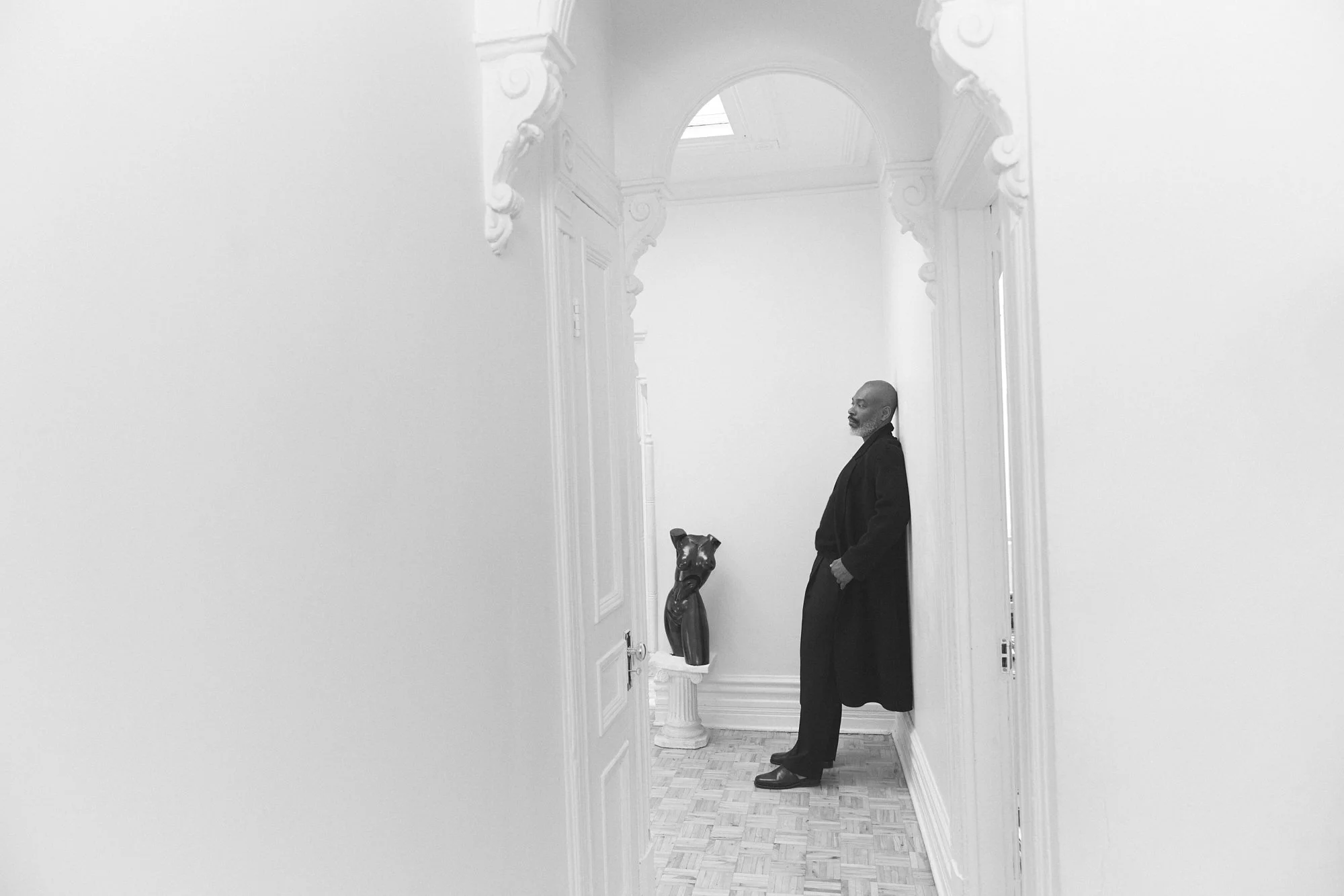 Un homme debout dans un couloir blanc avec un plafond voûté, une sculpture de chien sur une colonne à gauche, en noir et blanc.