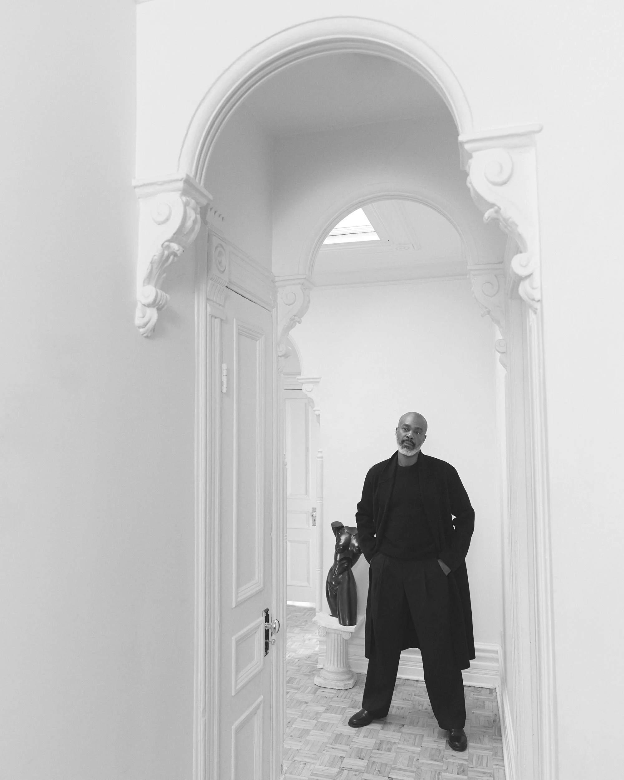 Homme noir debout dans une pièce élégante avec décoration classique, photographié à travers une porte ornée.