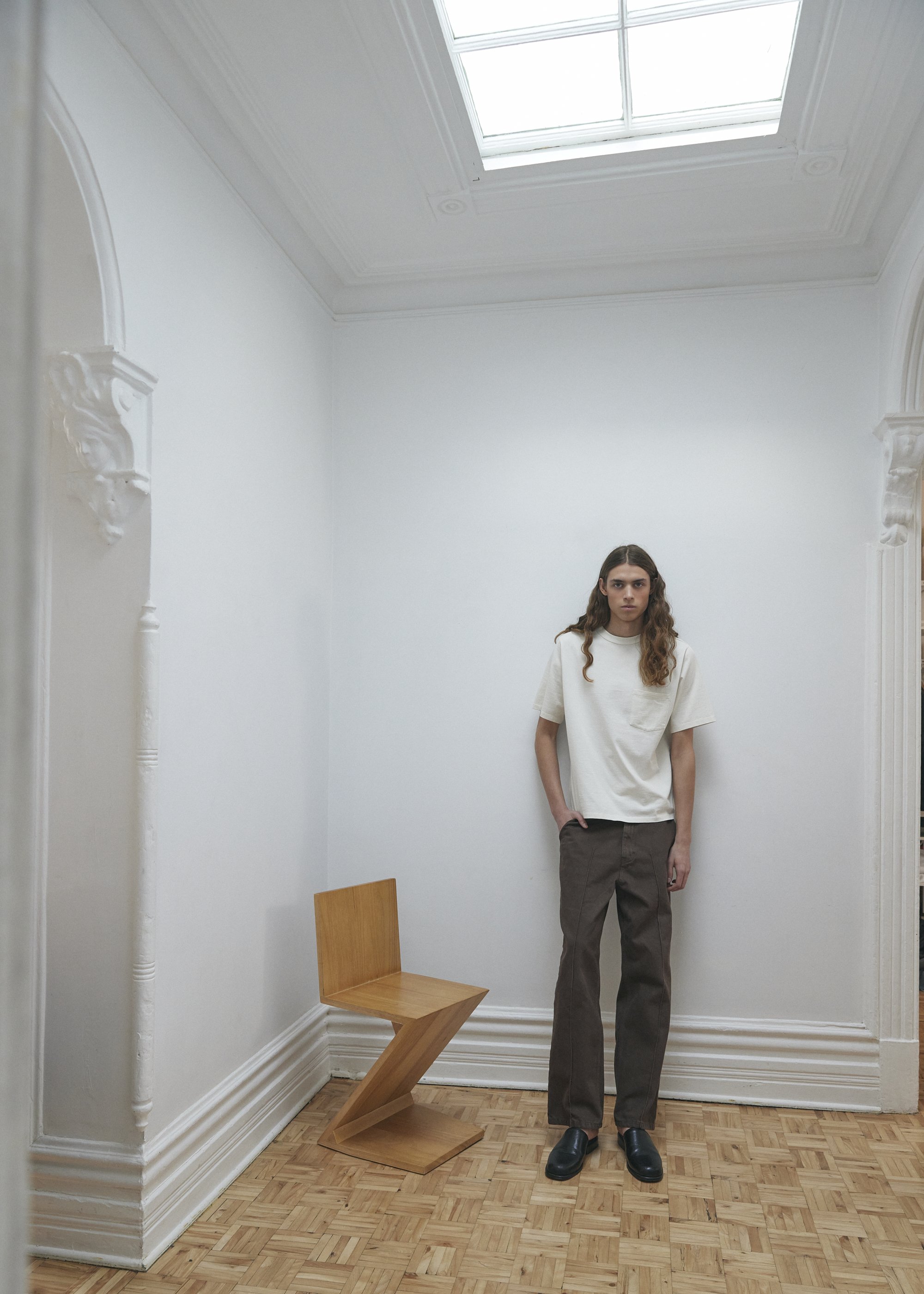 Une femme se tient dans une pièce blanche avec un plafond en verre, une chaise en bois stylisée et des décorations sculptées sur les murs.