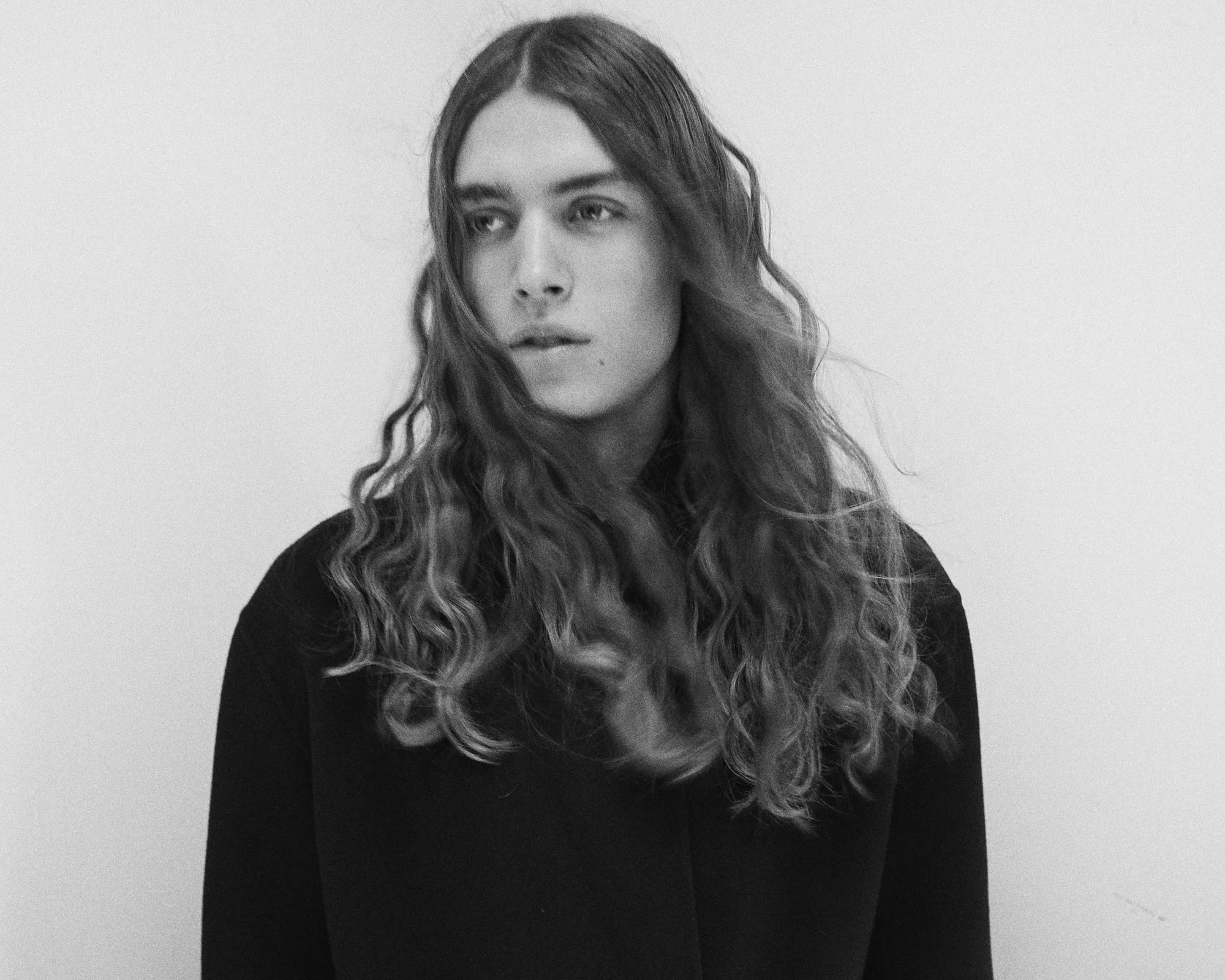 Jeune homme aux longs cheveux ondulés, portant un vêtement noir, regardant à gauche dans un cadre monochrome.