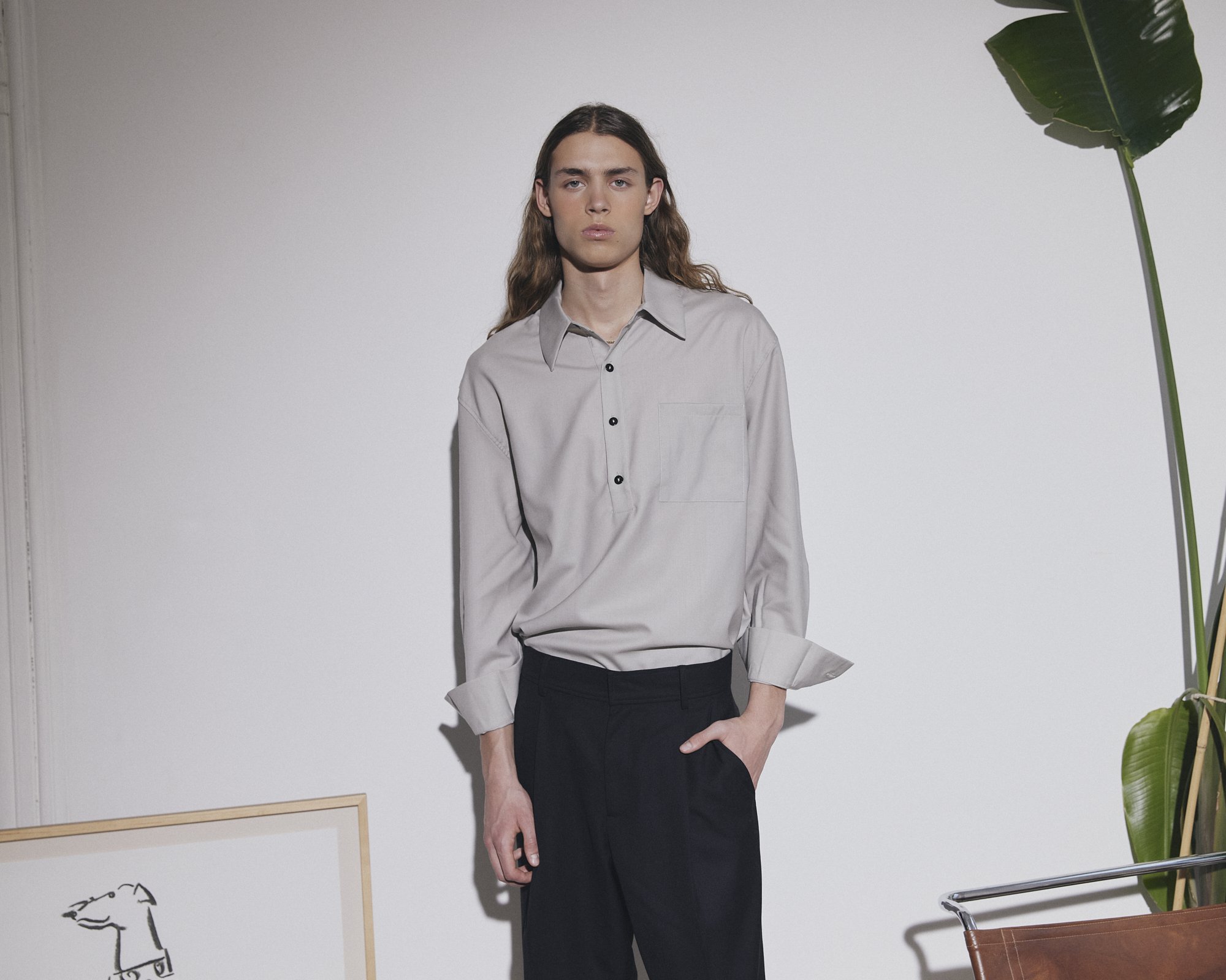 Jeune homme portant une chemise grise à manches longues et un pantalon noir, debout dans une pièce au mur blanc, avec une grande plante verte à droite et un tableau ou une affiche à gauche
