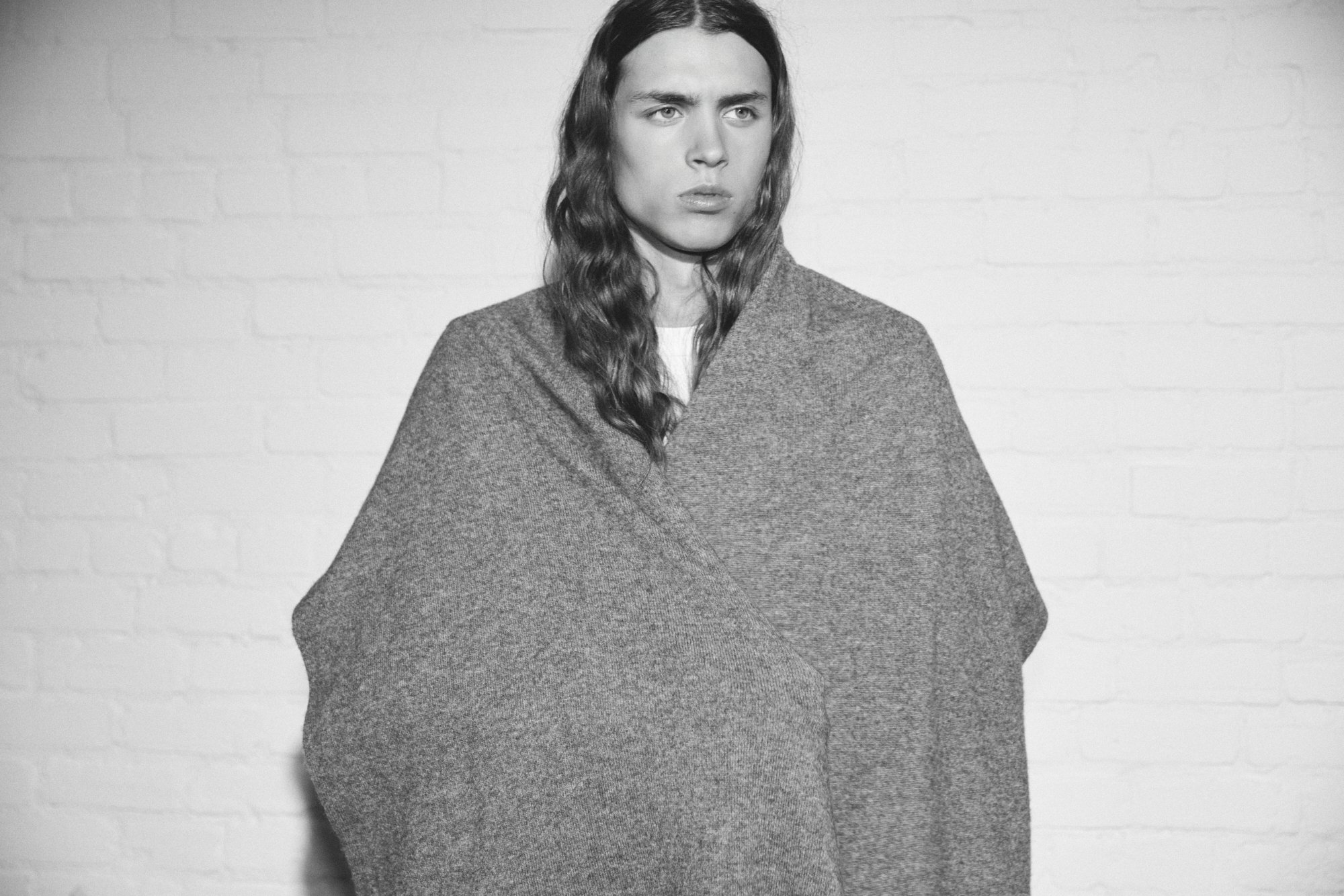 Une femme avec des cheveux longs, portant une cape ou un manteau gris, se tient devant un mur blanc en briques, dans une photo en noir et blanc.