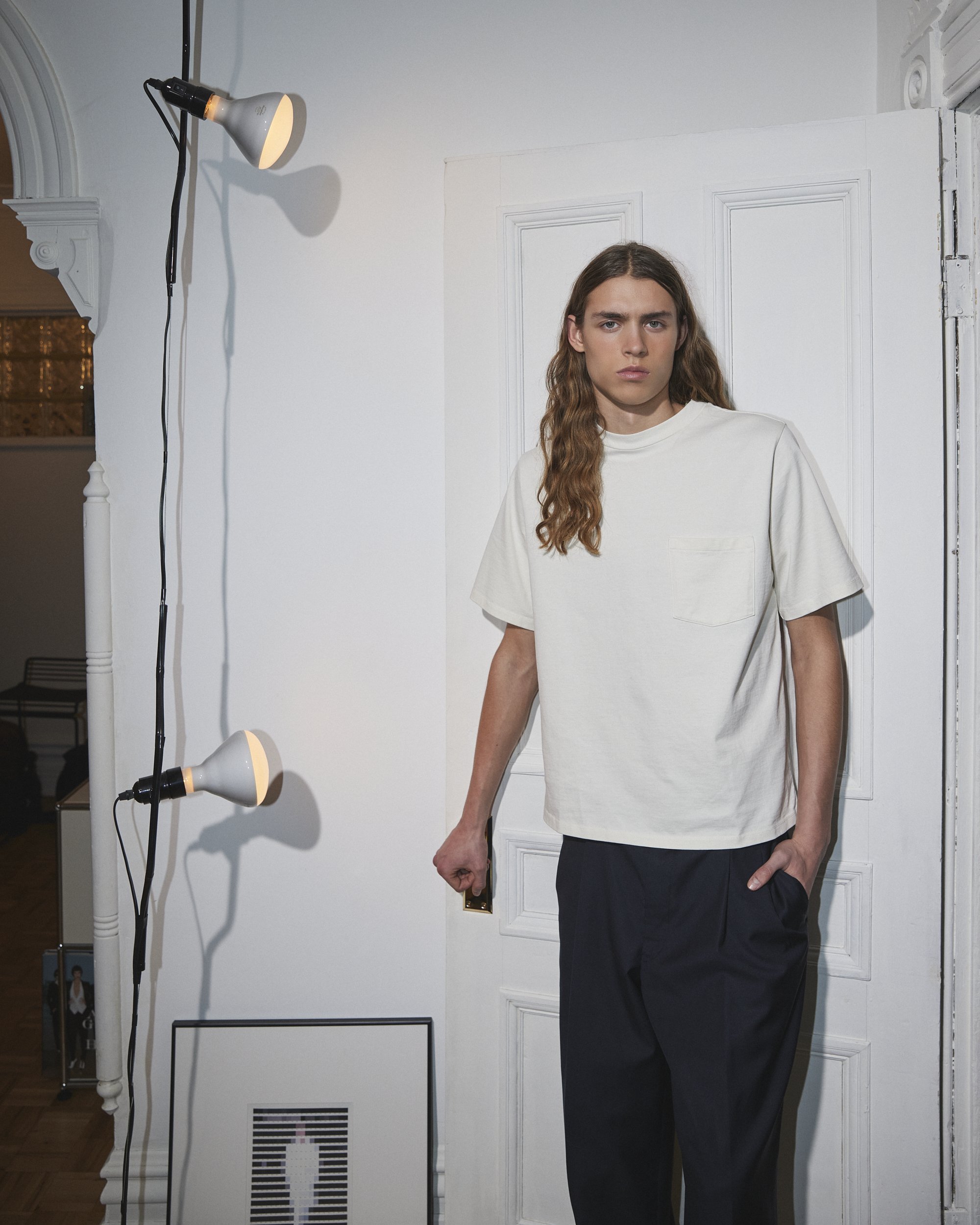 Jeune homme avec longs cheveux, portant un t-shirt blanc et un pantalon noir, debout dans une pièce lumineuse, avec un éclairage professionnel à gauche et un fond blanc.