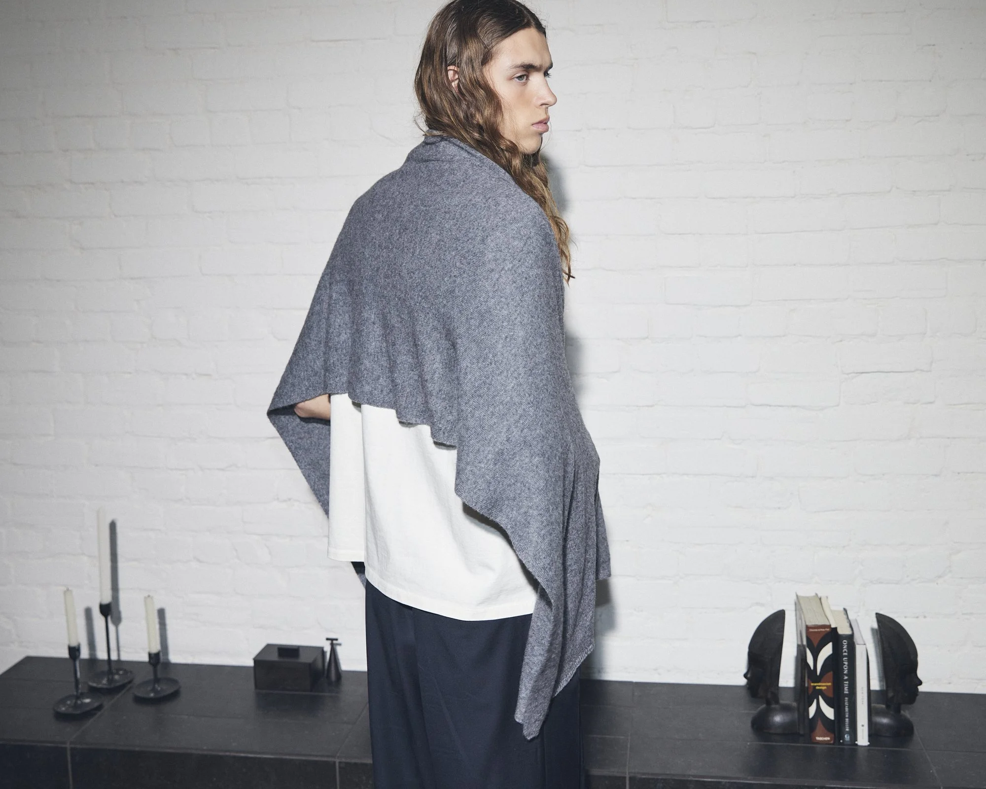 Une femme avec des cheveux longs et bruns pose contre un mur blanc en briques. Elle porte un vêtement gris avec une cape ou un châle et un pantalon foncé. Sur une surface en bas à gauche, il y a des bougeoirs avec des bougies, et à droite, une petite