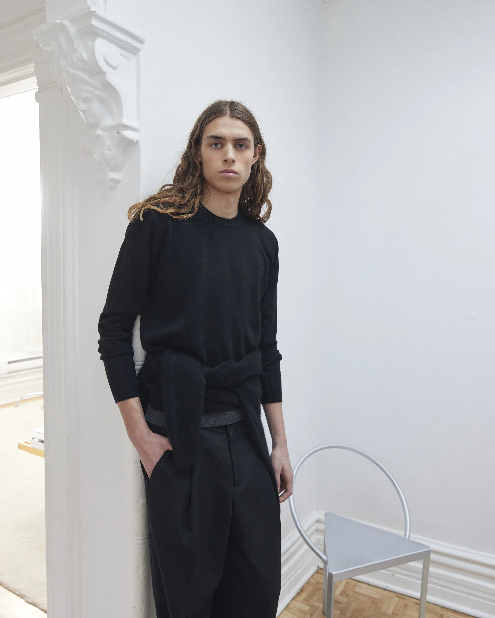 Jeune homme aux cheveux longs et raides portant un pull noir, debout dans une pièce blanche, à côté d'un mur décoré d'une moulure élégante. Il a une posture détendue avec une main dans la poche.