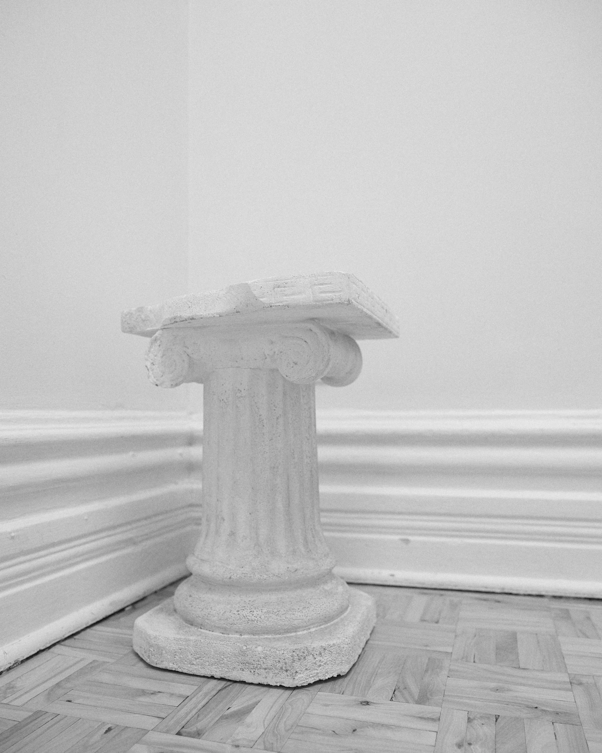 Statue en forme de colonne avec une plaque gravée en haut, en pierre, dans une pièce avec moulures en bois blanc et parquet en bois.