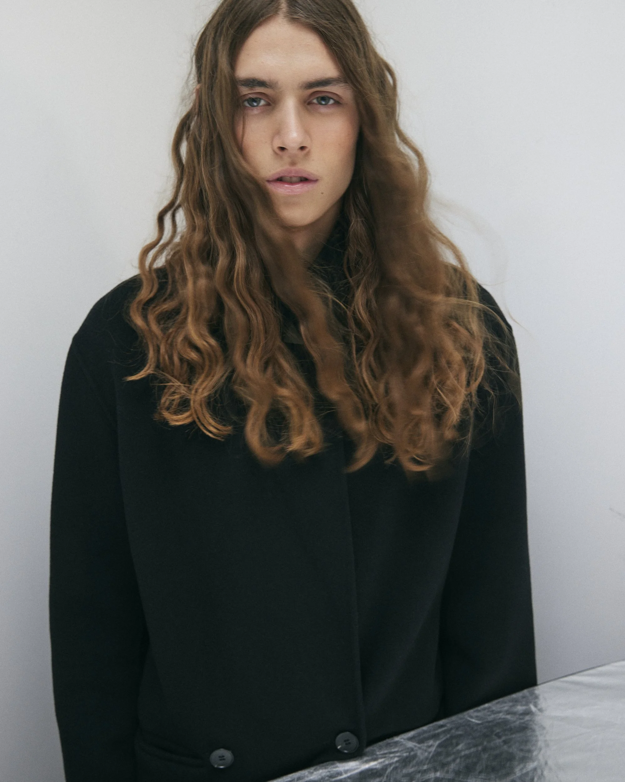 Jeune femme aux longs cheveux bouclés, portant un blazer noir, assise devant une table en marbre, regardant l'objectif. 
