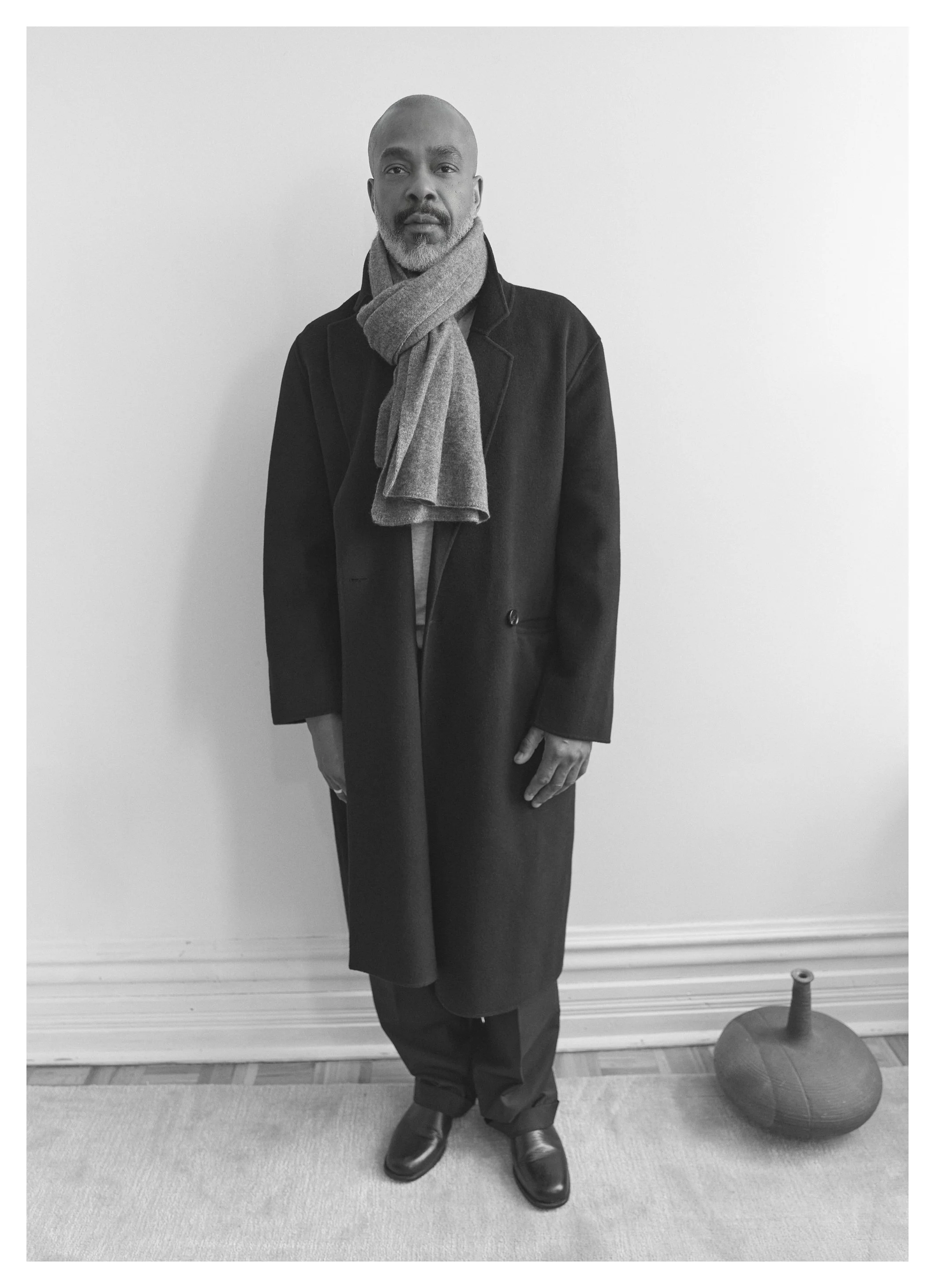 Un homme debout devant un mur blanc, portant un manteau long et un foulard, dans une pièce avec un tapis et un objet décoratif à côté de lui.