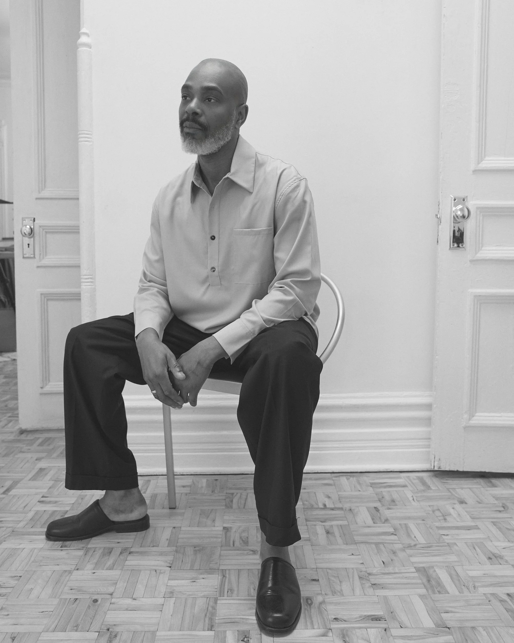 Homme assis sur une chaise dans une pièce vide, en noir et blanc, portant une chemise à boutons et des chaussures noires.