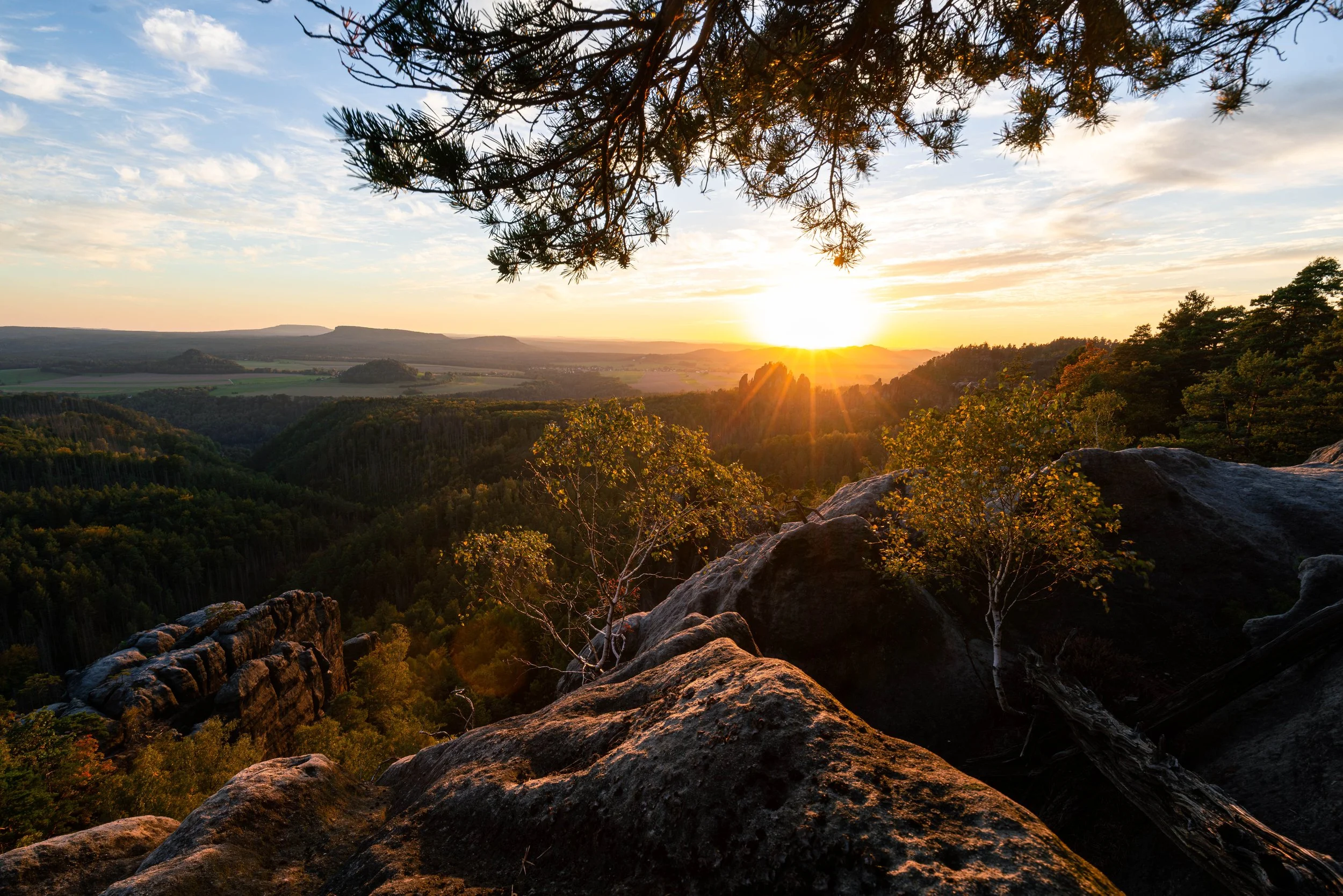 Sonnenuntergang über einer bewaldeten Landschaft mit Felsen im Vordergrund, einige Wolken am Himmel, sichtbare Baumäste im oberen Bereich.
