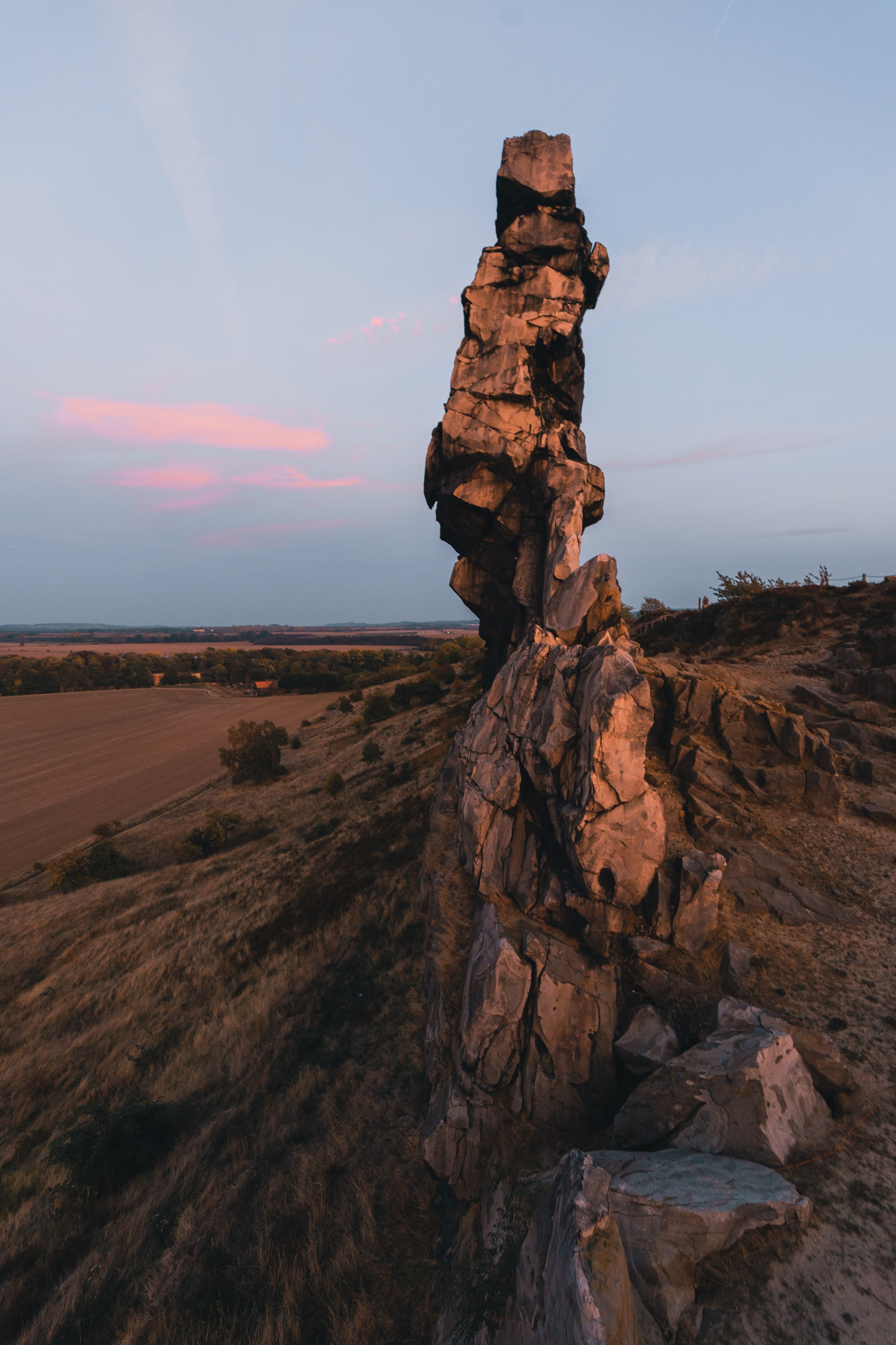 Ein hoher, geologischer Felsen im ländlichen Gebiet bei Sonnenuntergang, umgeben von Feldern und leicht bewaldeten Flächen im Hintergrund.