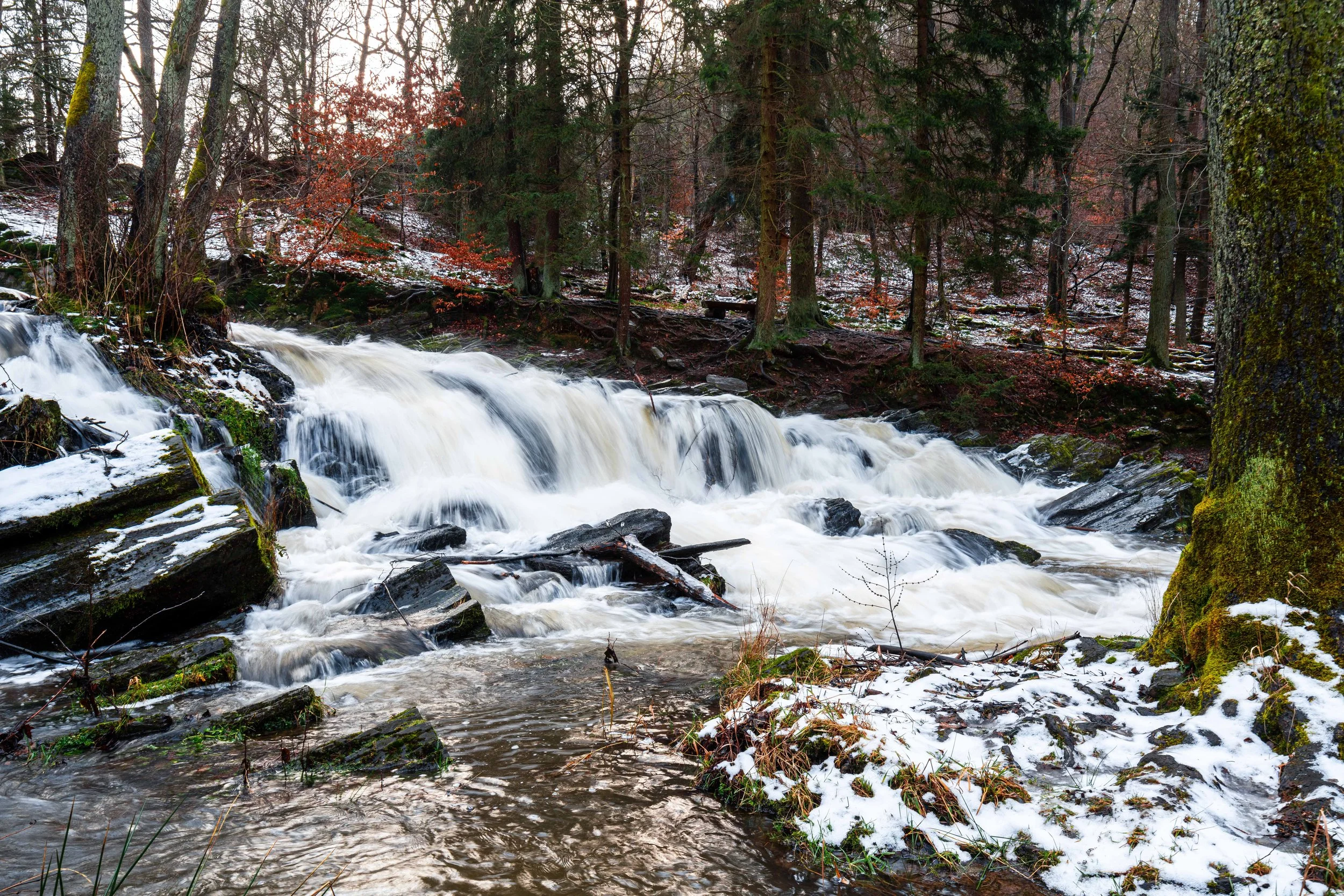 Ein Wasserfall im Wald mit Bäumen, Schneeresten und Felsen.