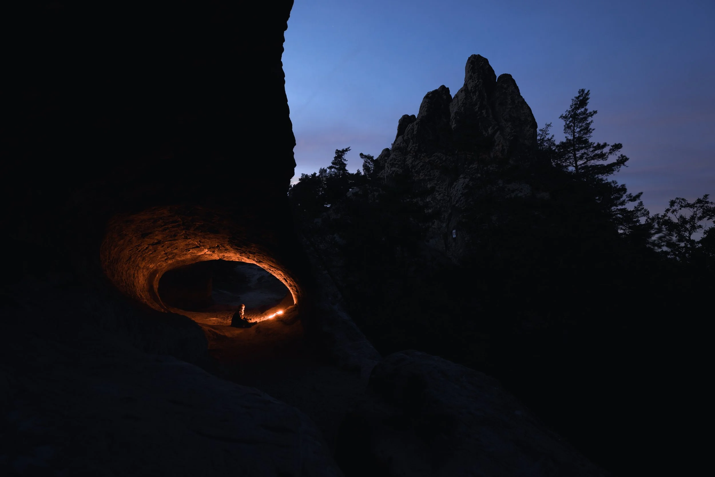 Eine Höhle bei Nacht mit einem Feuer oder Kerze im Inneren. Draußen sind Bäume und Felsen vor einem bläulichen Himmel sichtbar.