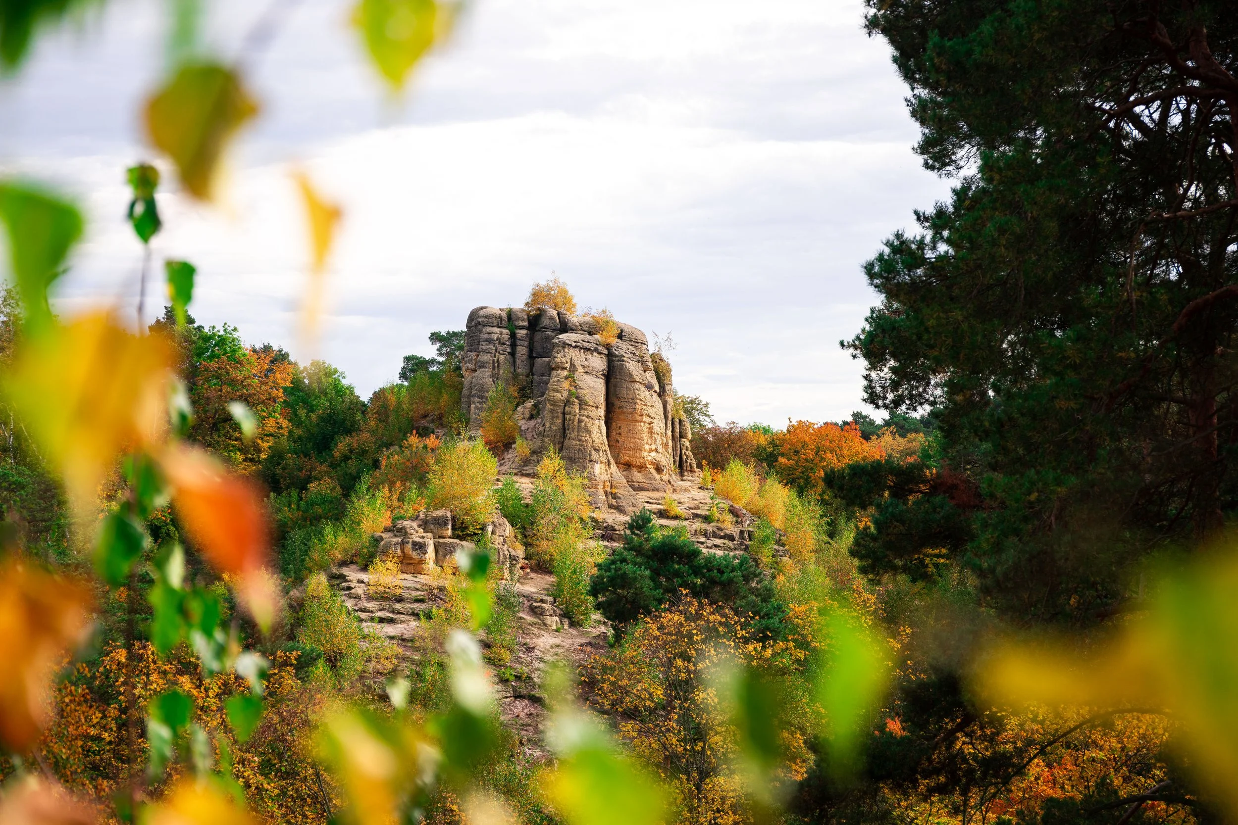 Felsformation auf einem Hügel inmitten eines Waldes mit herbstlicher Laubbedeckung, umgeben von bunten Bäumen in orange, gelb und grün, mit einem bewölkten Himmel im Hintergrund.