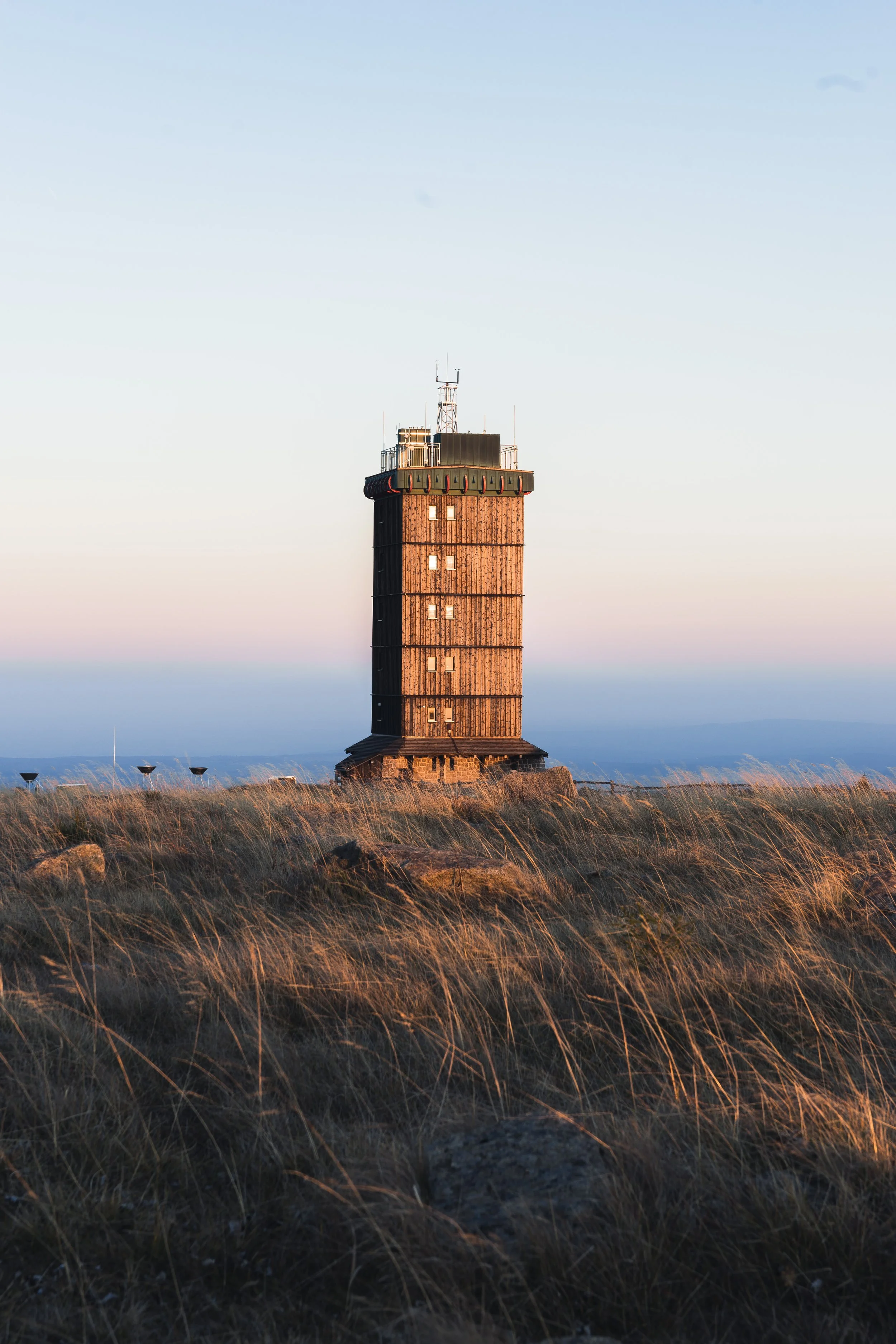 Ein hoher, rechteckiger Leuchtturm aus Holz auf einer felsigen, grasbewachsenen Hügellandschaft bei Sonnenuntergang.